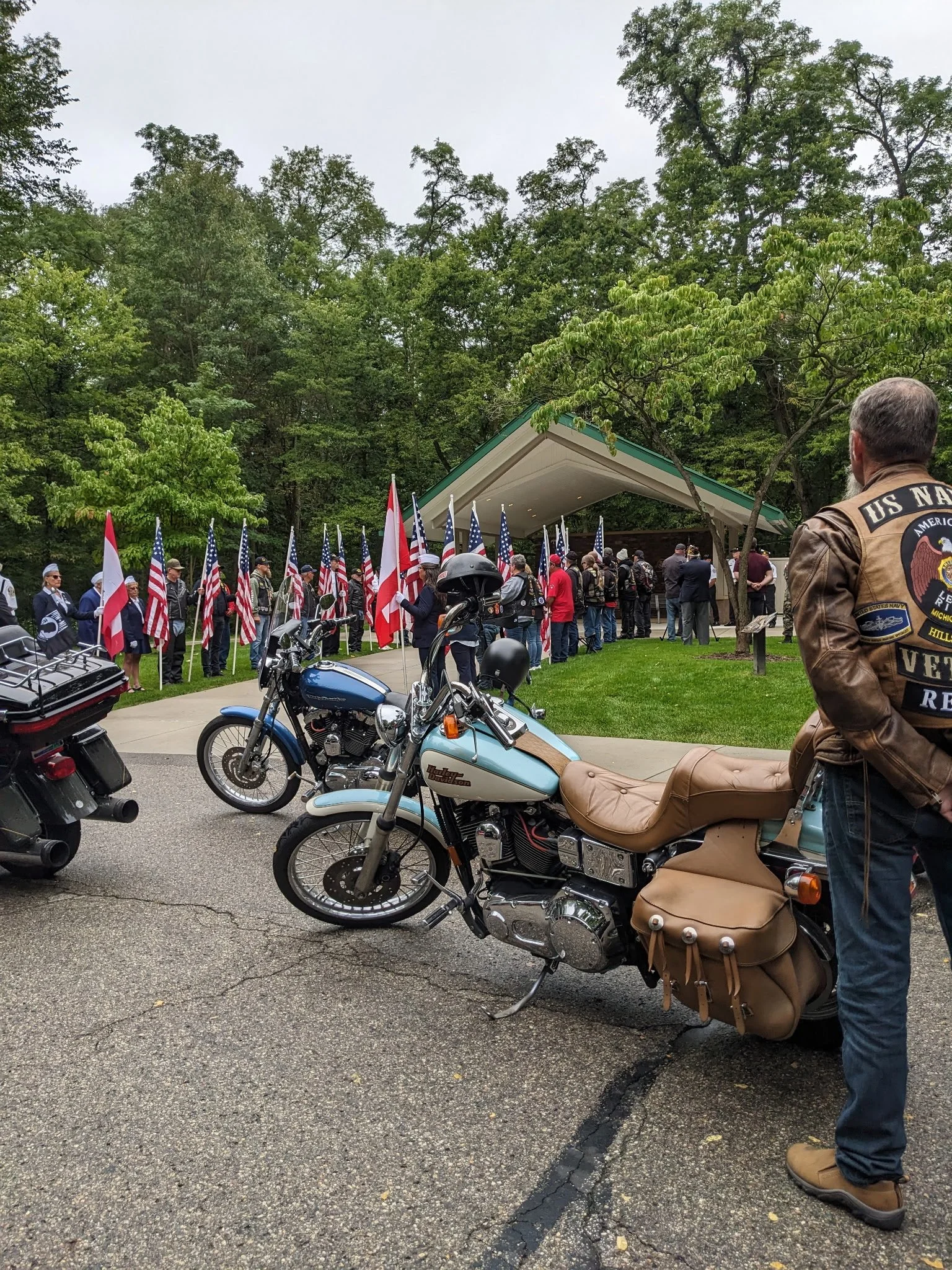 People gather at an outdoor memorial with flags, including American flags, and motorcycles parked nearby. One man wears a leather patch vest with U.S. Navy emblem.