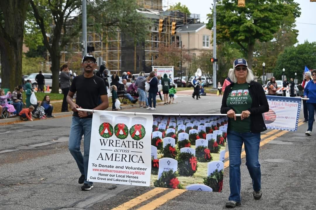 Two people walking in a parade holding a banner that reads "WREATHS across AMERICA" and features images of grave markers decorated with wreaths. The parade takes place on a city street with spectators, some seated and some standing, watching from the sidewalk. There are trees, houses, and a construction building in the background.
