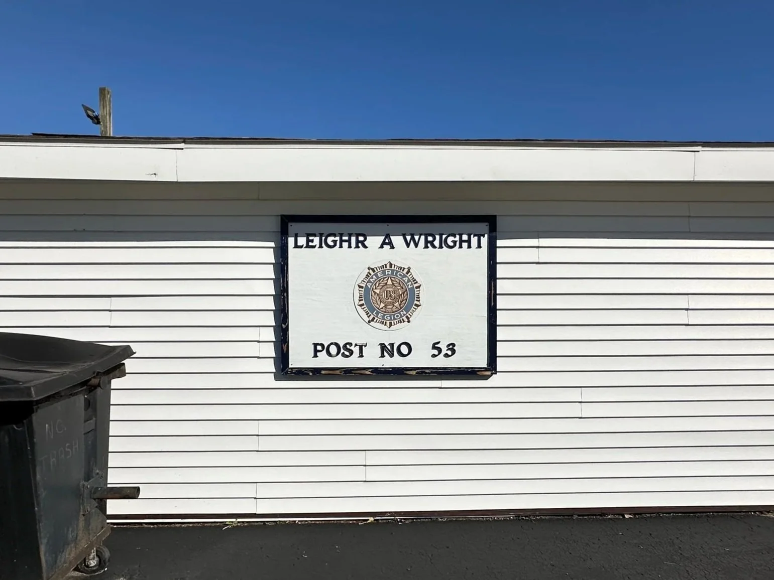 Sign for Leigh A. Wright Post 53 of the American Legion mounted on a white siding building, with a trash bin in the foreground and a clear blue sky above.