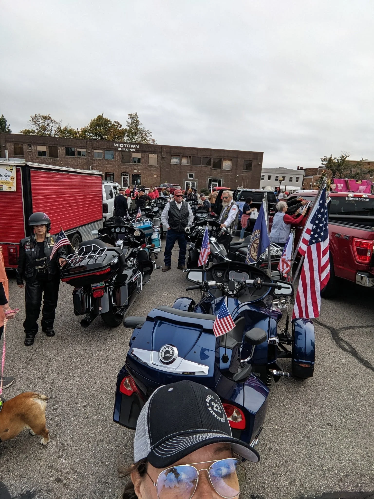 A gathering of people with motorcycles and flags in a parking lot in front of a brick building, with a woman wearing sunglasses and a cap in the foreground.