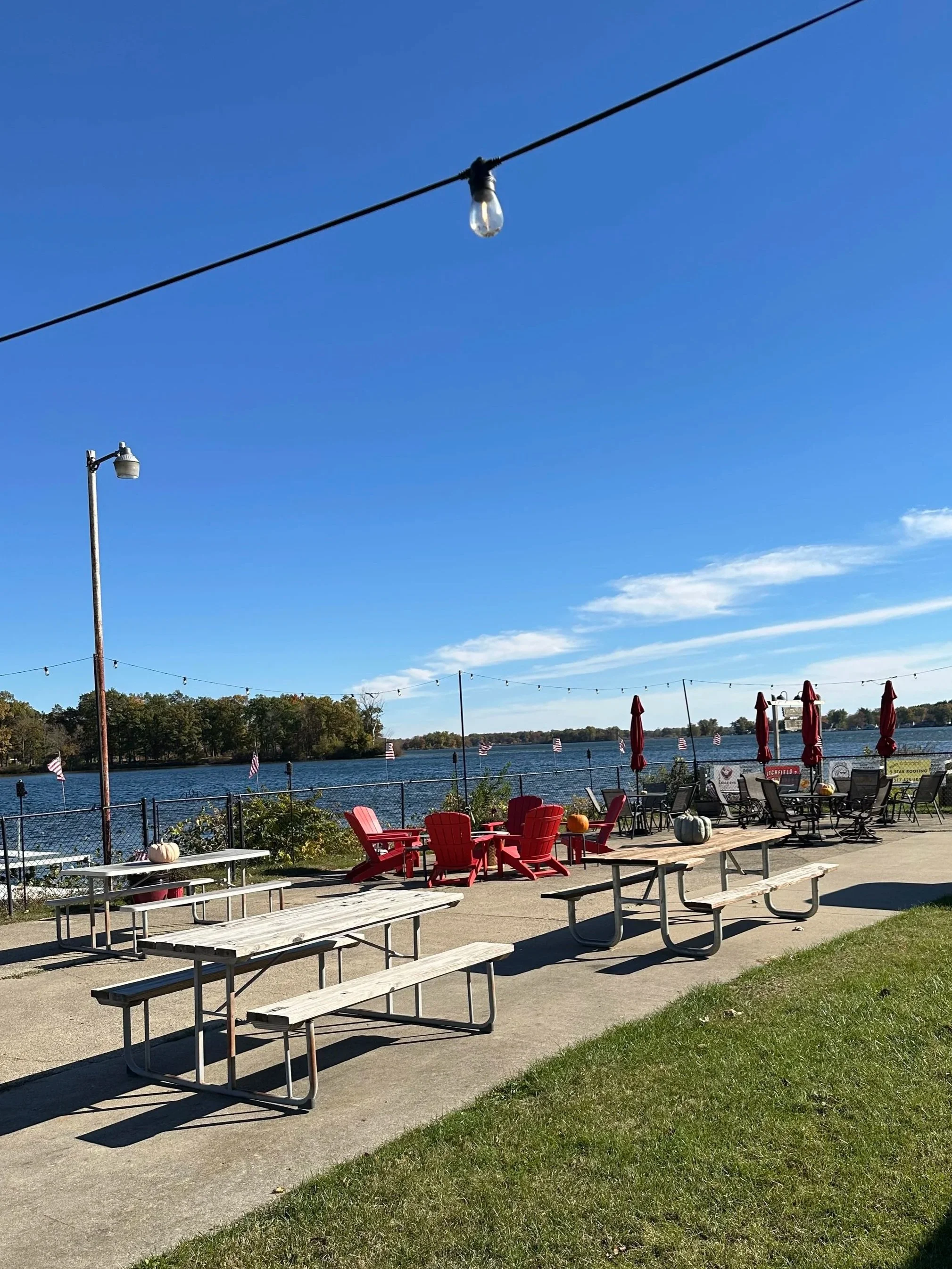 Outdoor patio area with picnic tables, Adirondack chairs, umbrellas, pumpkins, and string lights by a body of water on a sunny day.