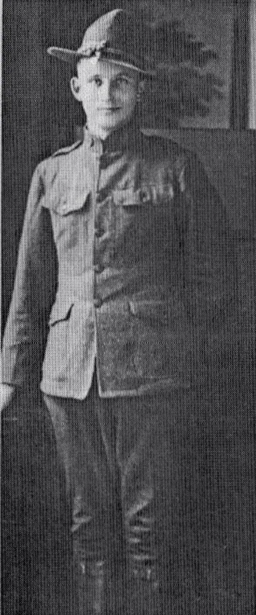 A young boy dressed in a vintage military uniform, wearing a wide-brimmed hat, standing indoors against a plain wall.