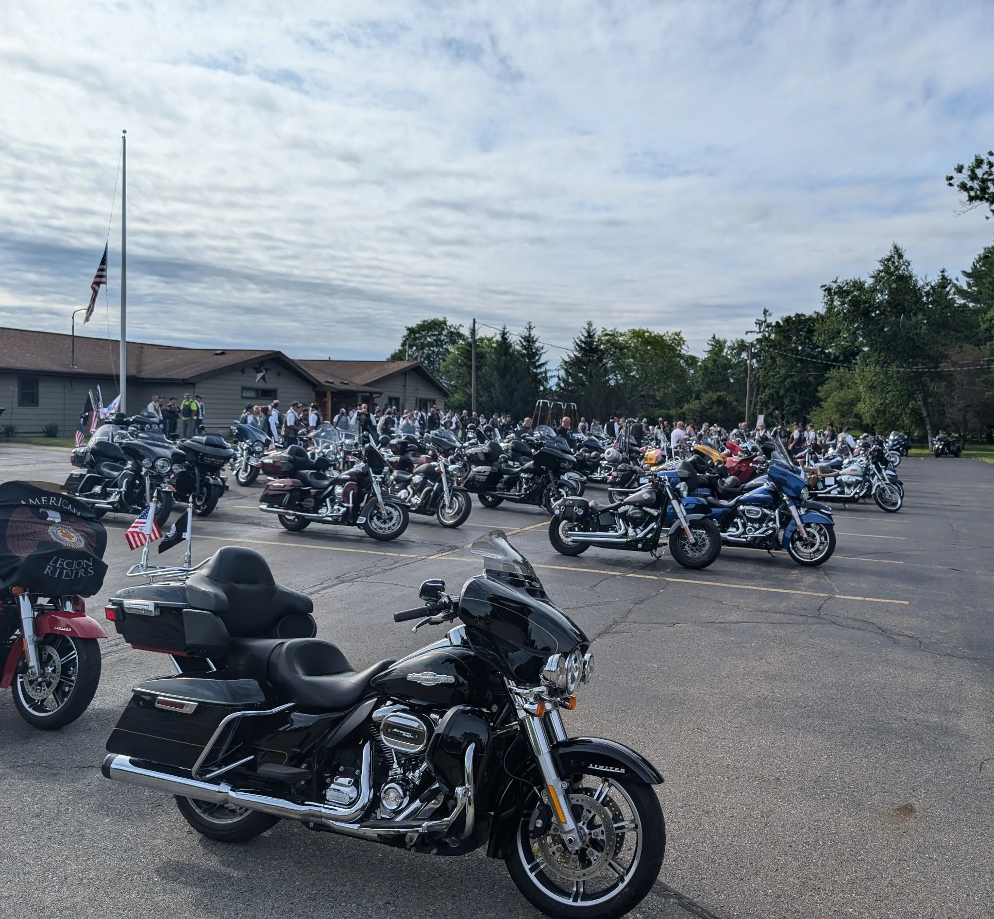 Black touring motorcycle parked in front of a gathering of motorcycles and people in a parking lot, with a building and trees in the background.