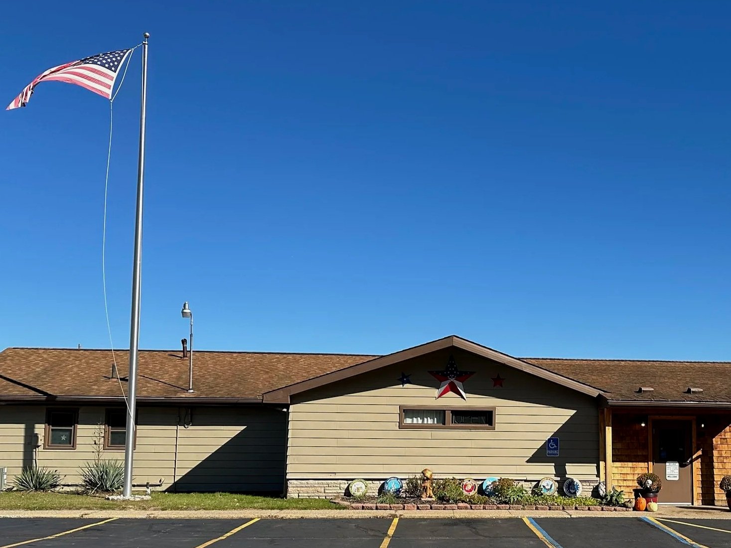 A single-story building with beige siding, decorated with red, white, and blue stars and patriotic items, with a large American flag flying on a flagpole in front, under a clear blue sky.
