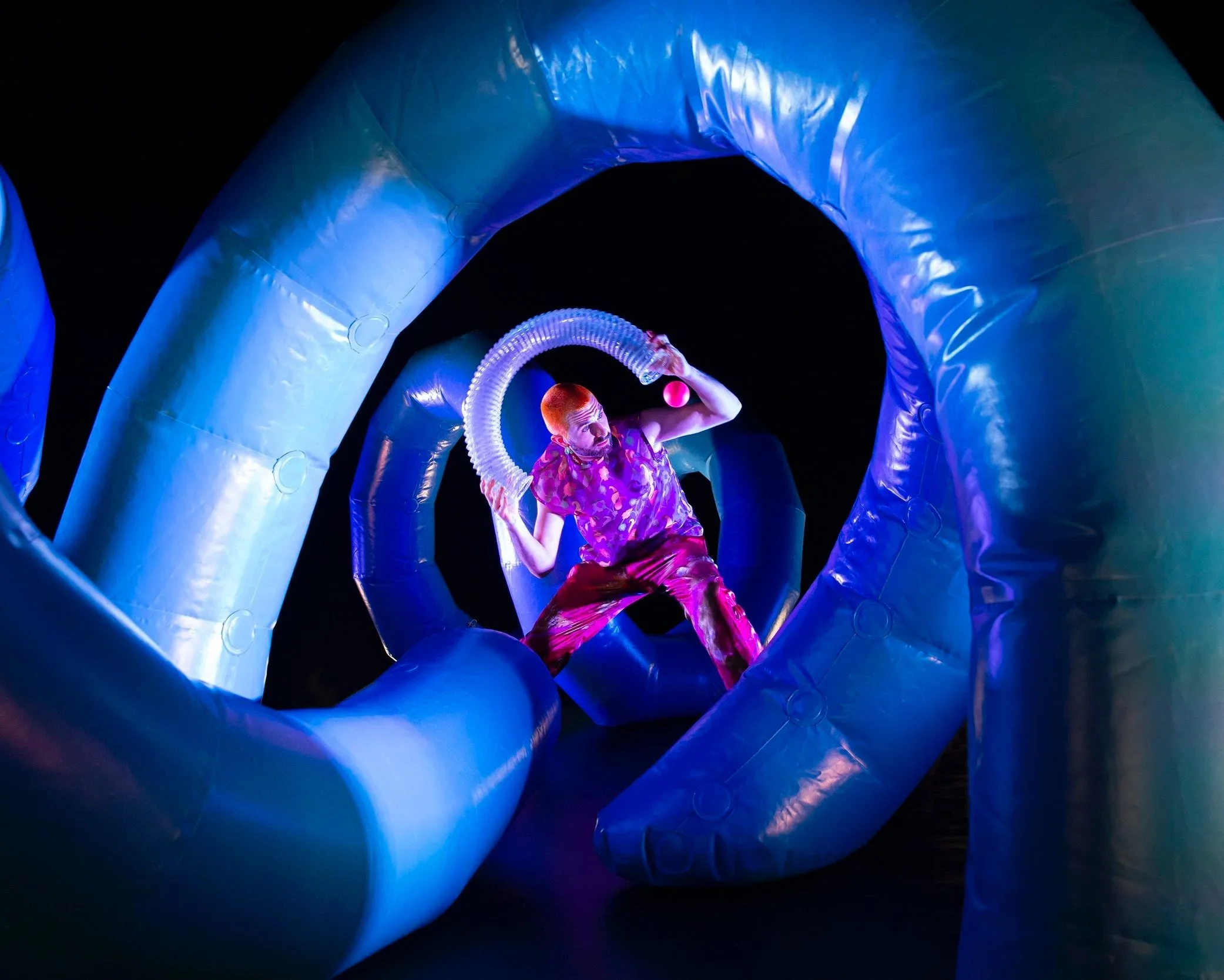 A performer with bright red hair, wearing a colorful purple and pink outfit, is surrounded by large inflatable blue tubes at night, illuminated by vibrant blue and purple lights.