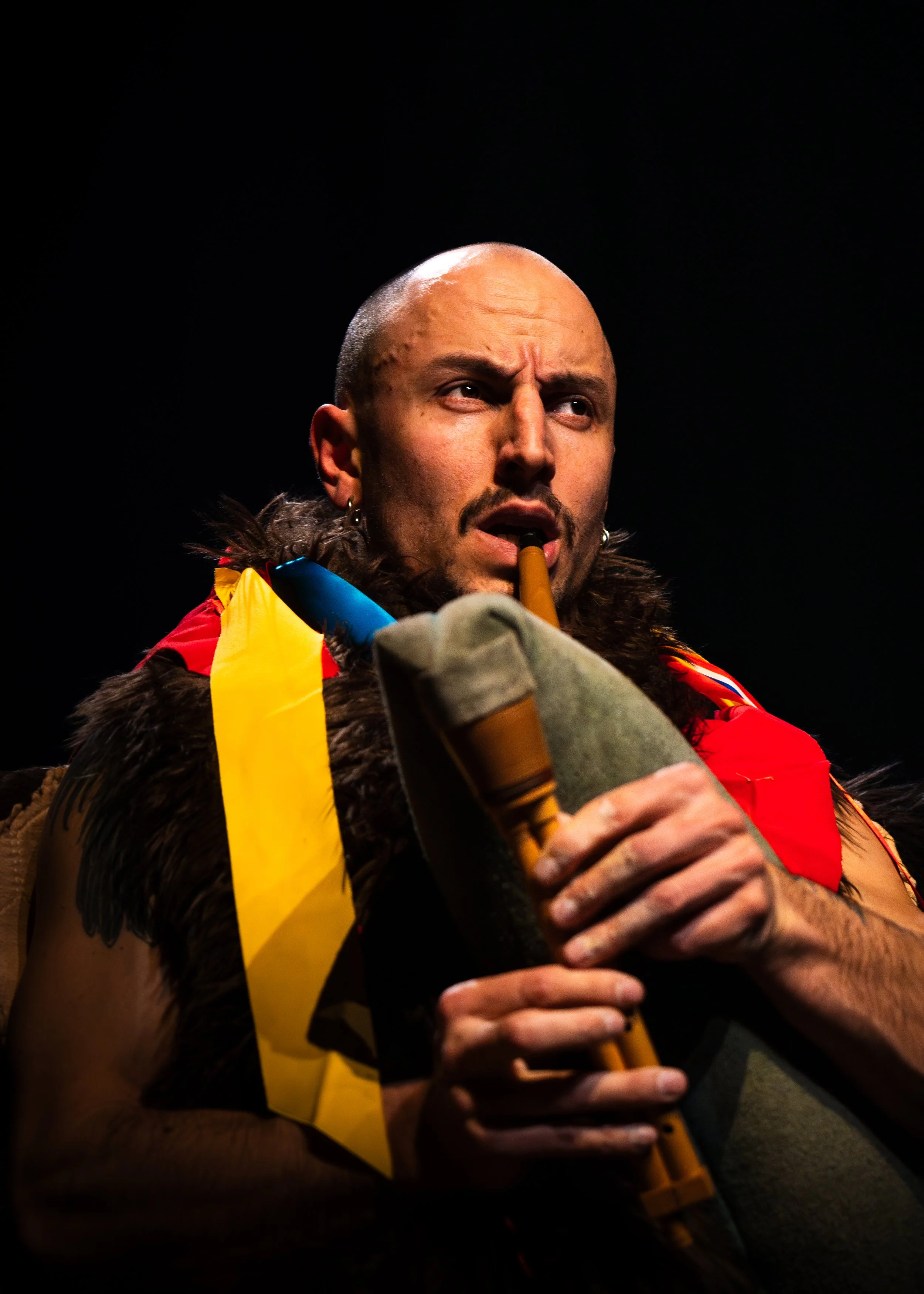 A person playing a didgeridoo, wearing a red garment, fur, and a yellow sash, with a dark background.