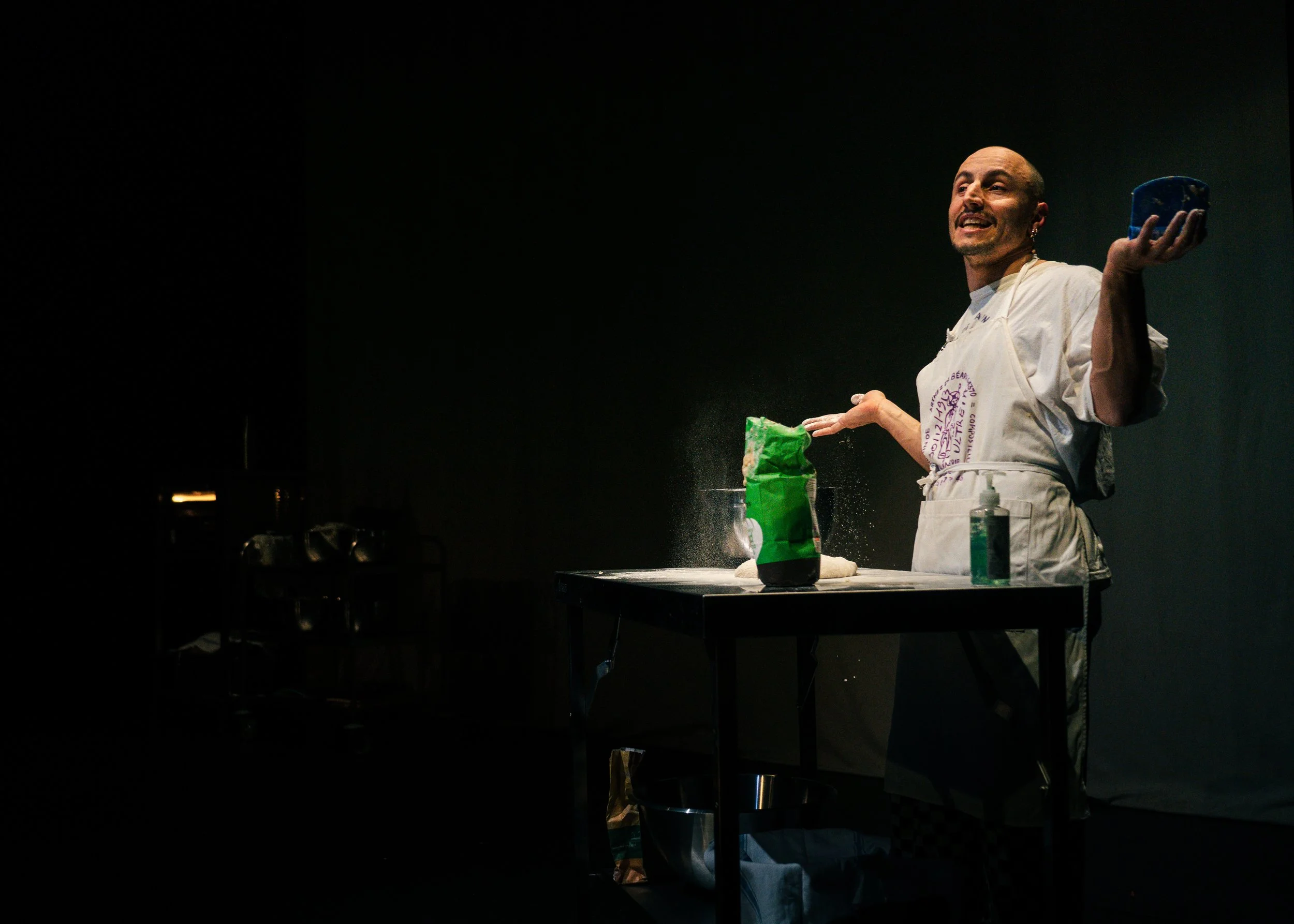 A person in a white apron and earrings is performing a cooking demonstration, holding a blue container in one hand and a green bag of ingredients on a table with a dark background.