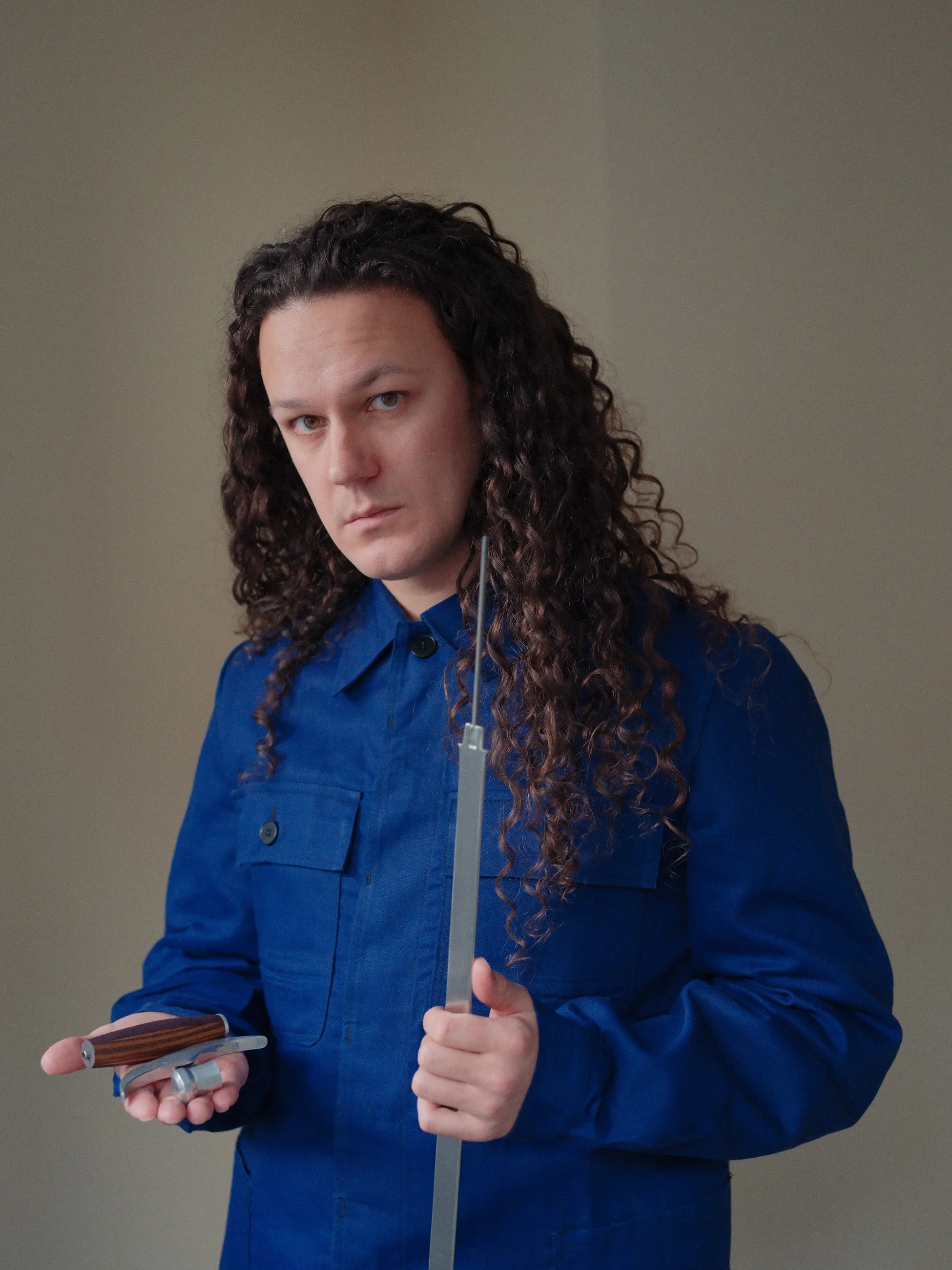 A man with long, curly dark hair wearing a blue work jumpsuit, holding a tuning fork and a small metal object, looking directly at the camera with a serious expression.
