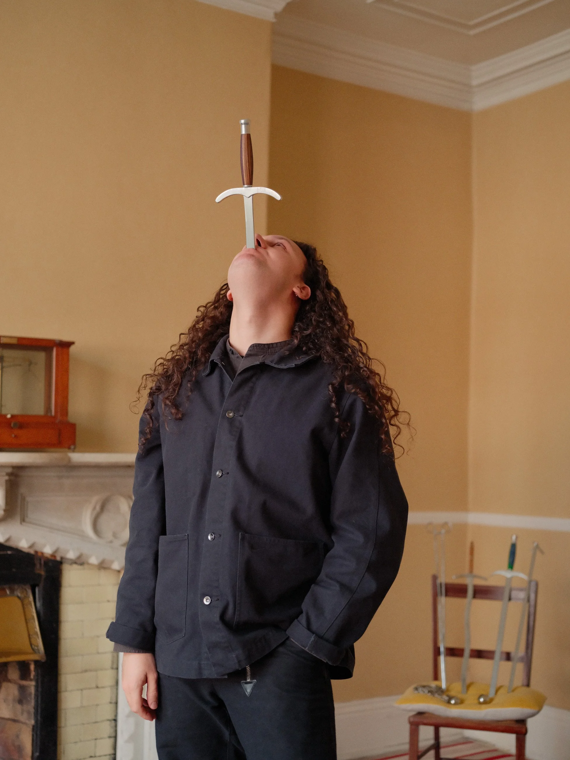 A person with long curly hair balancing a sword on their face in a room with yellow walls, a fireplace, and chairs with multiple swords stuck in a pillow on one of them.