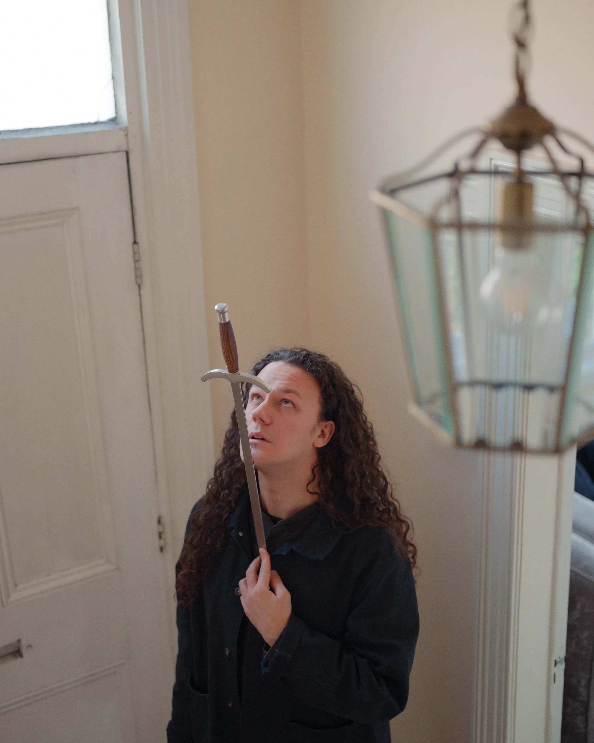 A person with long, curly hair holding a sword, looking up, standing near a door and a hanging lantern.