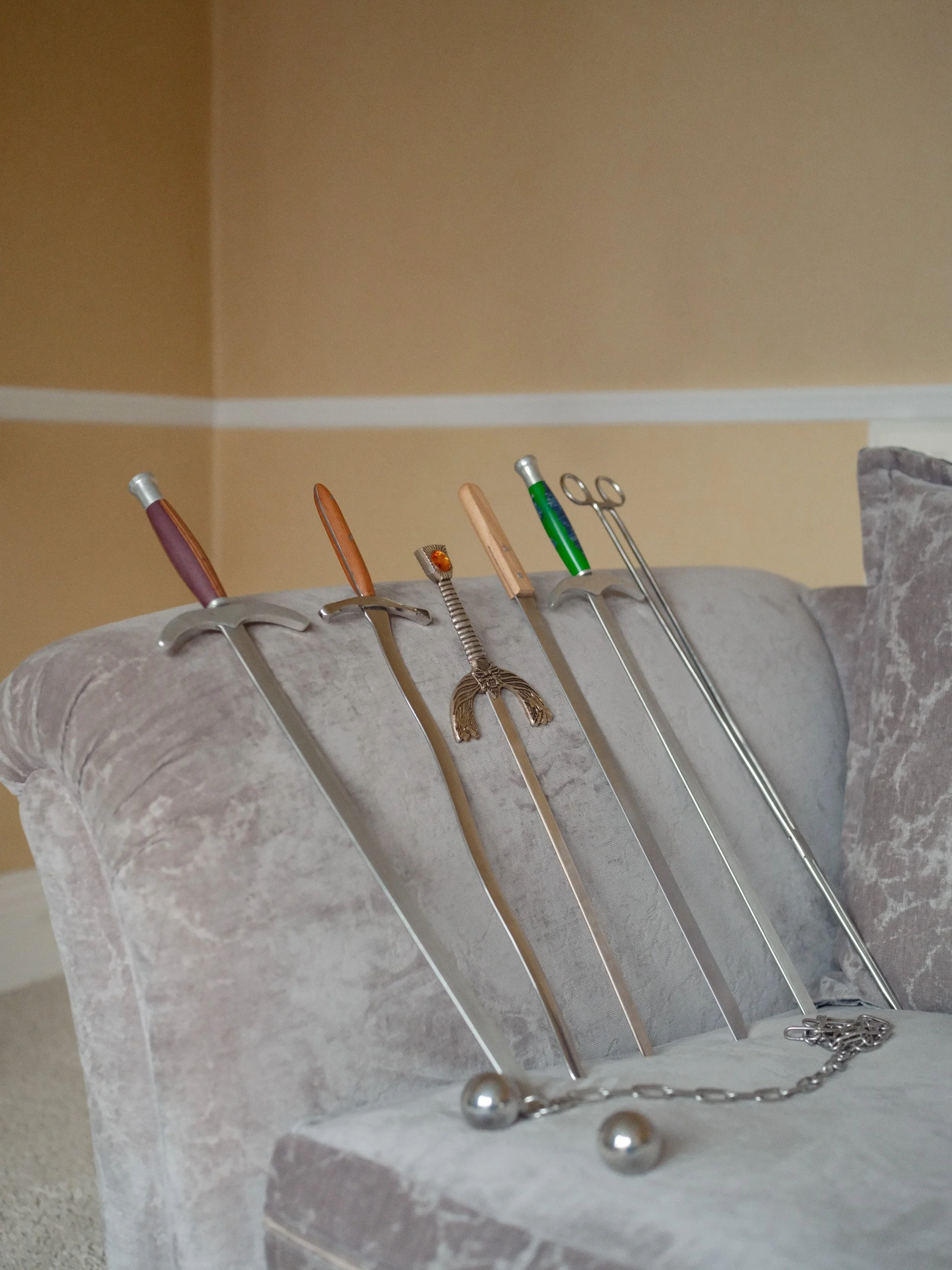 Collection of six miniature swords and a chain with a ball on each end, displayed on a plush beige armchair.