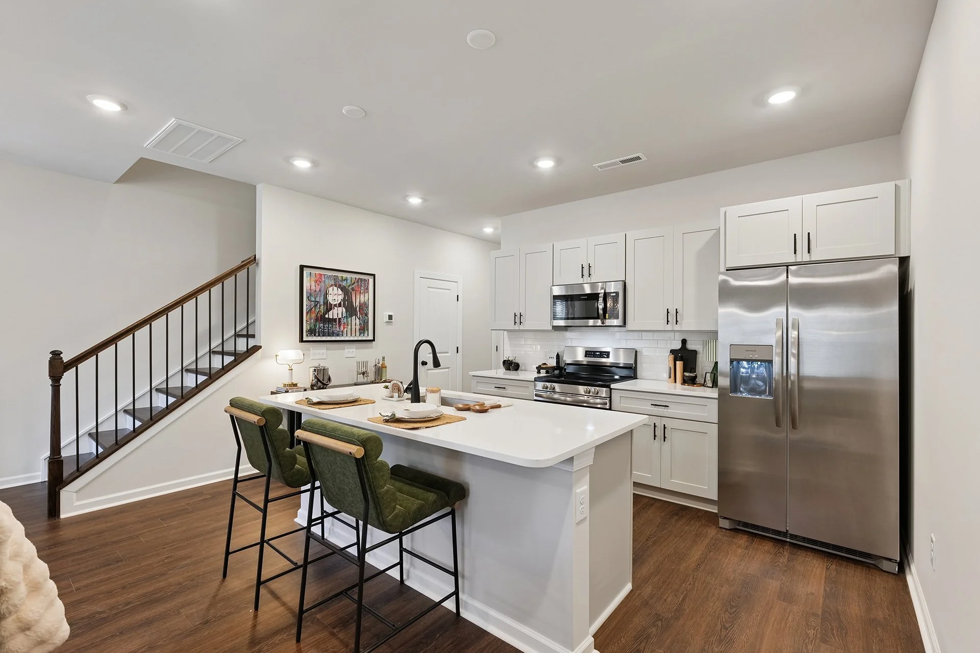 Modern kitchen with white cabinets, stainless steel refrigerator and oven, island with green chairs, wooden floor, and artwork on the wall.