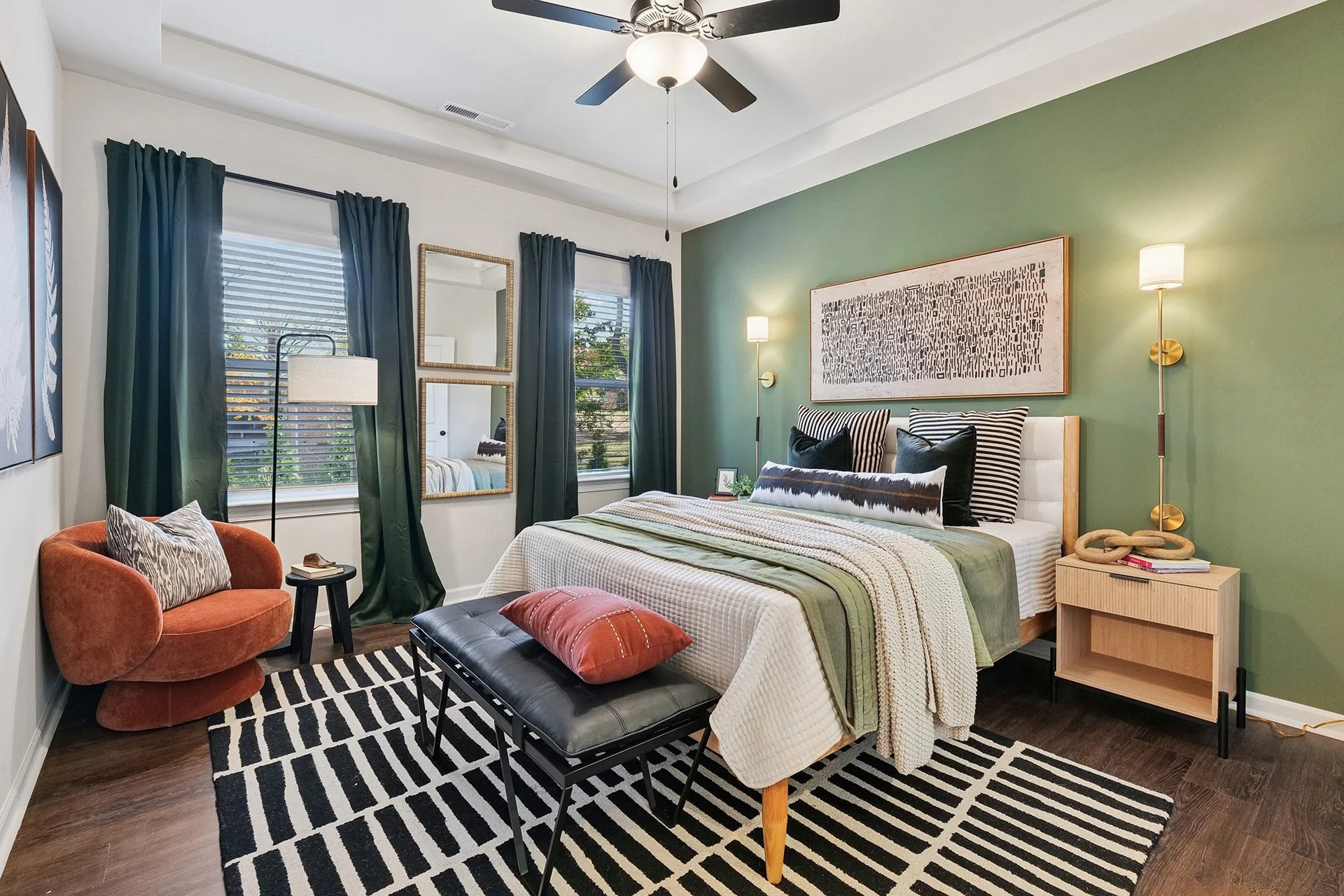 A modern bedroom with a green accent wall, a bed with patterned pillows, a black and white striped rug, an orange armchair, a black bench with a red pillow, and two windows with dark green curtains.