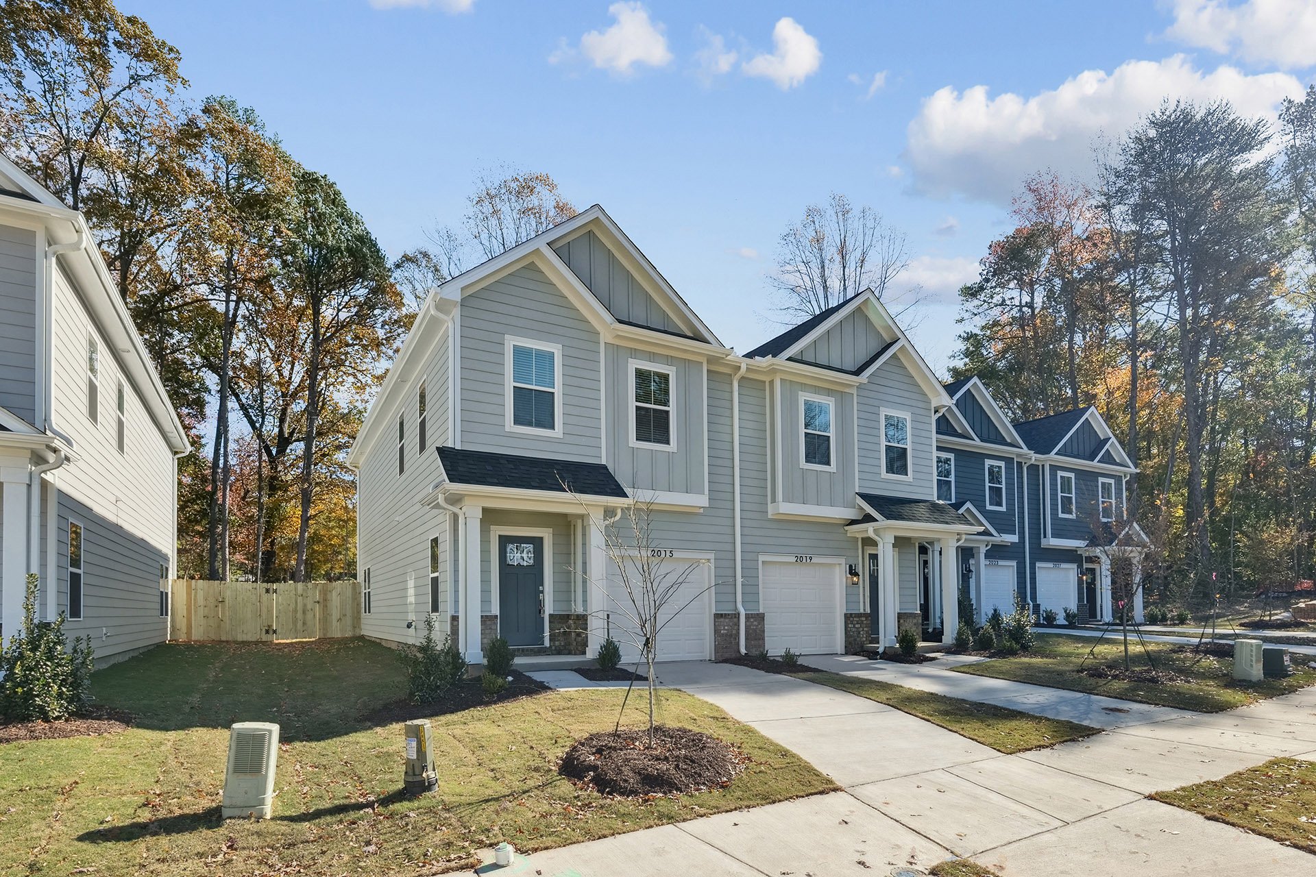Multiple newly built townhouses with front lawns, driveways, and a fence, under a partly cloudy sky, with trees in the background.