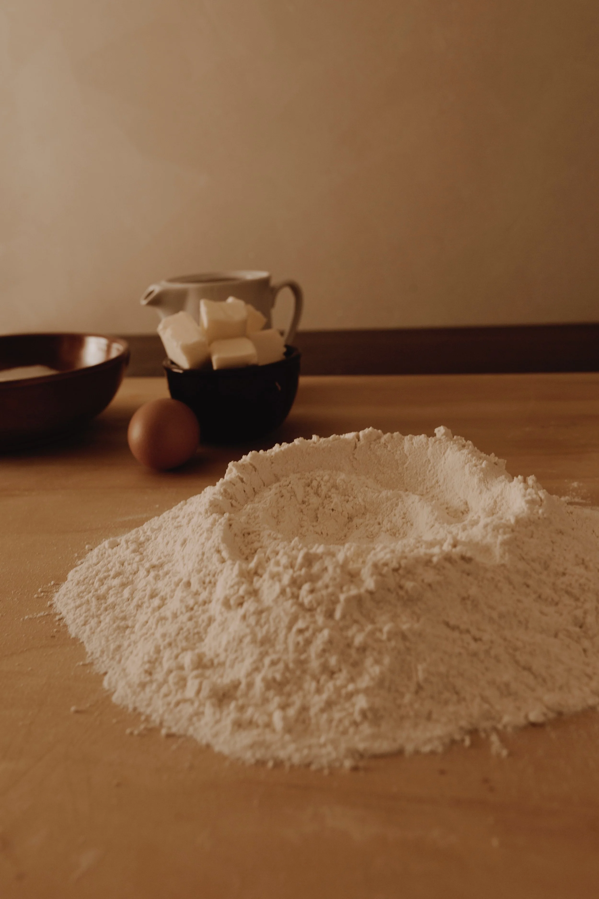 Harina en un montículo, un huevo, trozos de mantequilla, y tazas en una mesa de cocina, con fondo de madera.