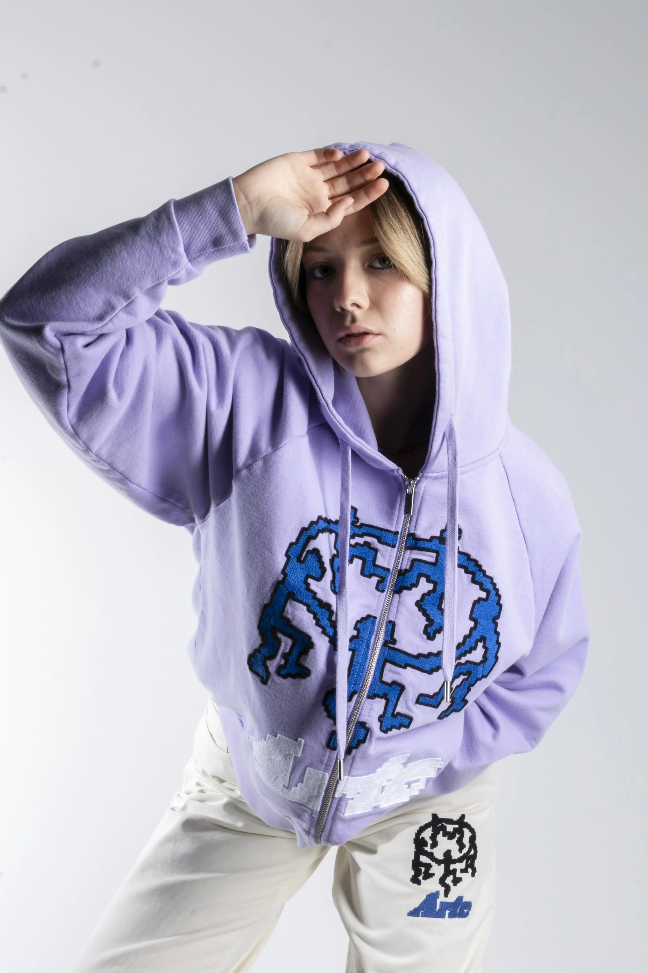 Jeune femme portant une veste à capuche violette avec un motif en pixel bleu et des pantalons blancs avec un patch noir et bleu, faisant un geste de la main pour saluer.