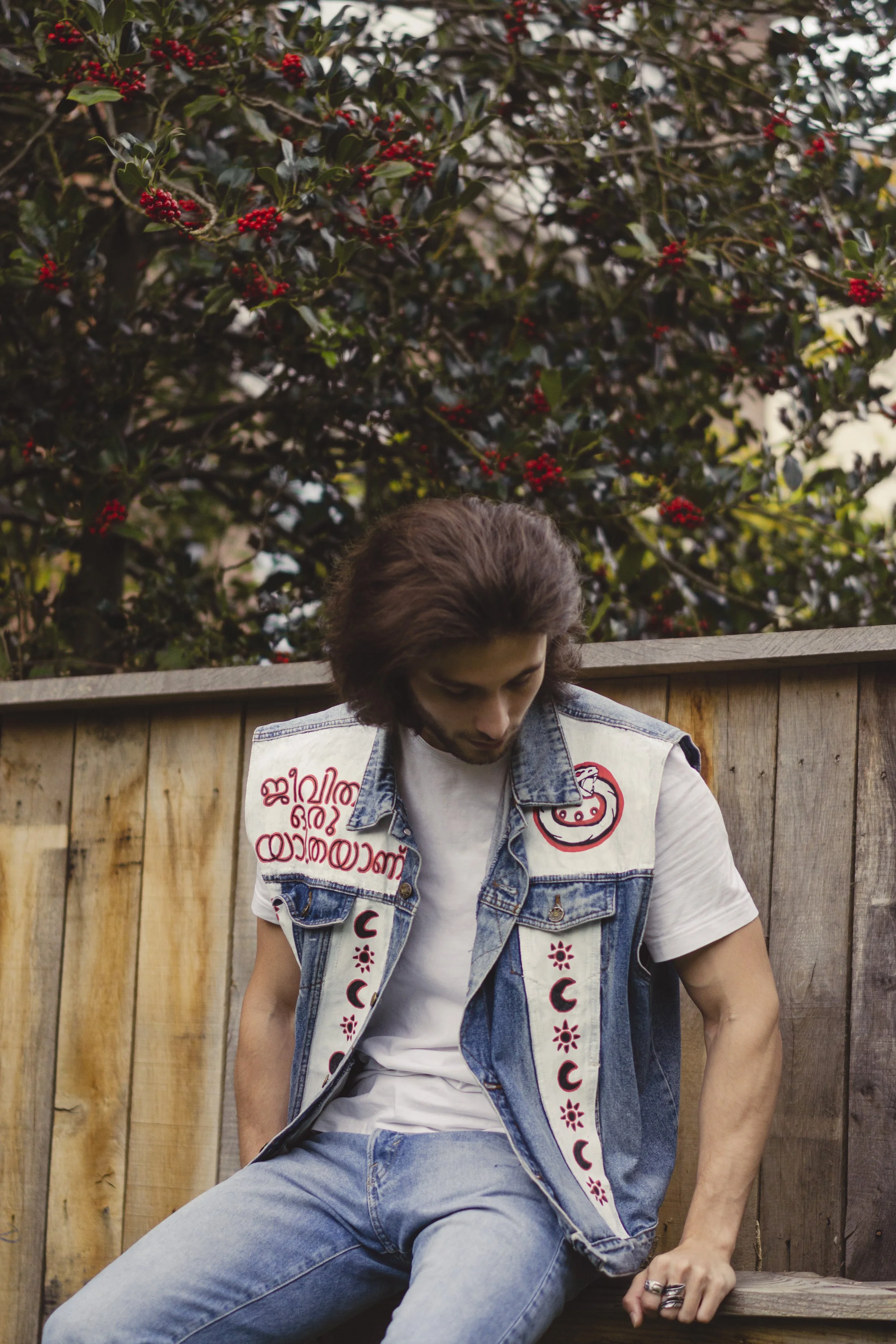 Jeune homme assis sur une clôture en bois, portant un gilet en jean avec des broderies rouges et blanches, un t-shirt blanc, et des jeans bleus, sous un arbre avec des baies rouges.