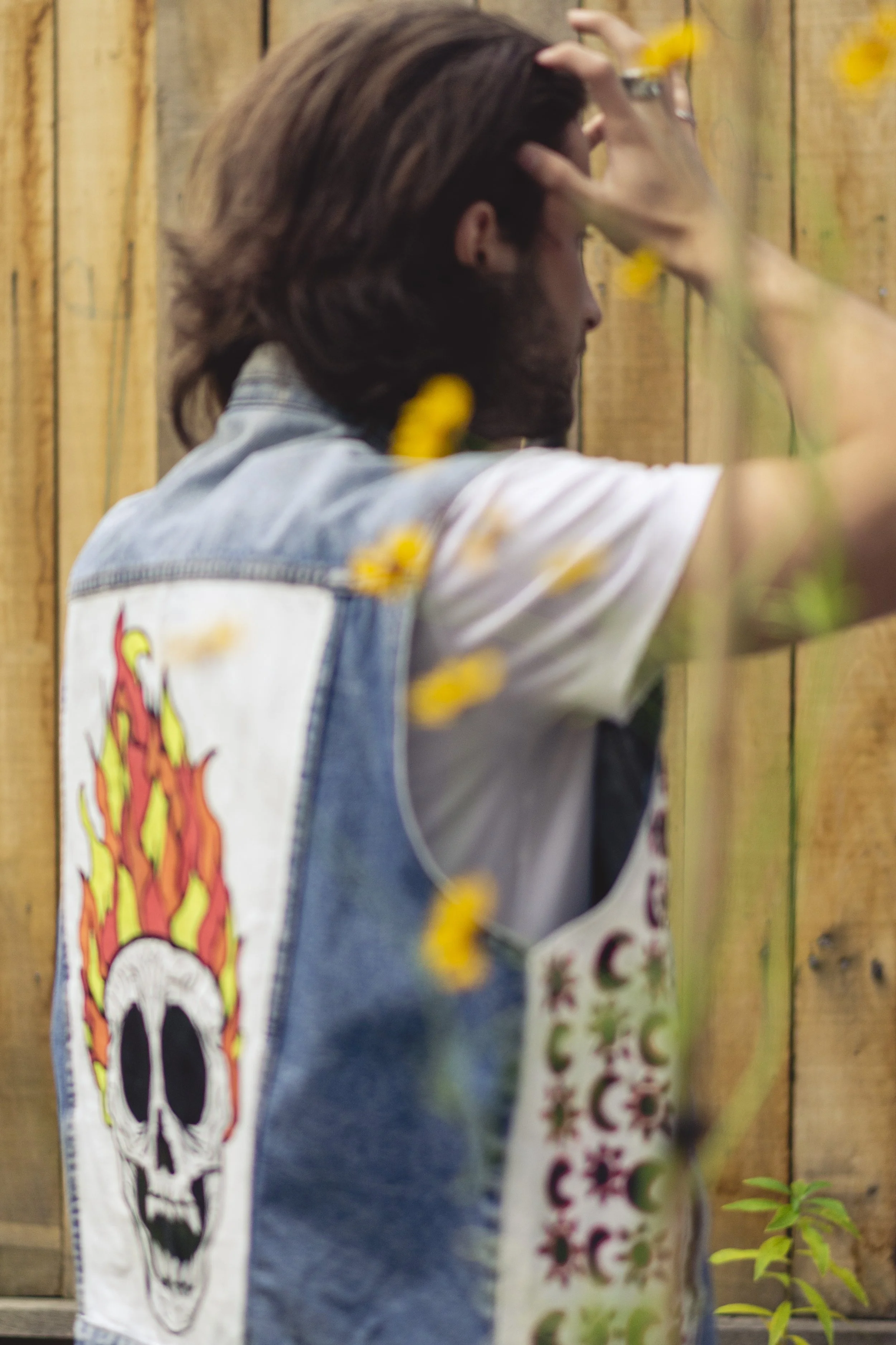 Personne avec les cheveux longs et barbe, portant un vêtement avec un patch de crâne en flammes, regardant vers un mur en bois, avec des fleurs jaunes en avant.