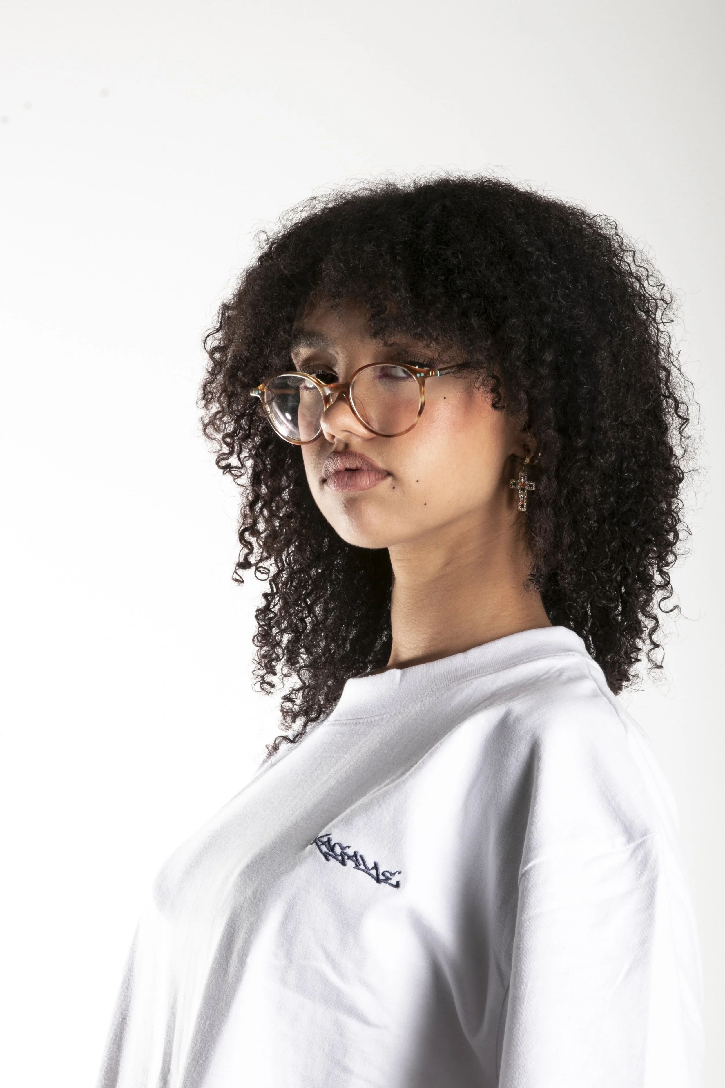 Jeune femme avec des cheveux bouclés noirs, porte des lunettes rondes, des boucles d'oreilles à croix, vêtue d'un t-shirt blanc avec un logo en haut à gauche.