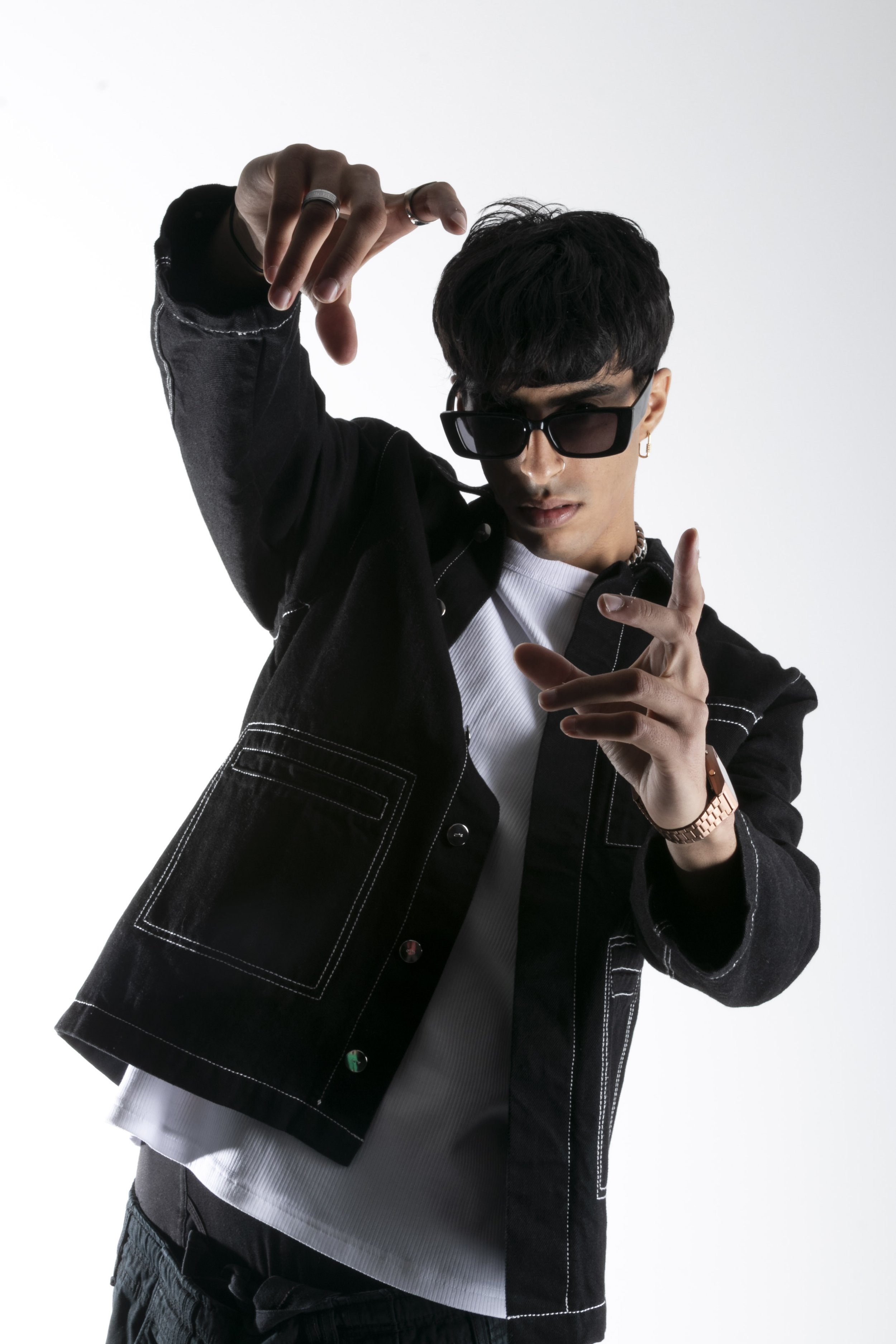 Jeune homme posant avec des lunettes de soleil noires, portant une veste en jean noire avec des coutures apparentes, un t-shirt blanc en dessous, un bracelet en or et une bague, faisant un geste de danse ou de pose dynamique.