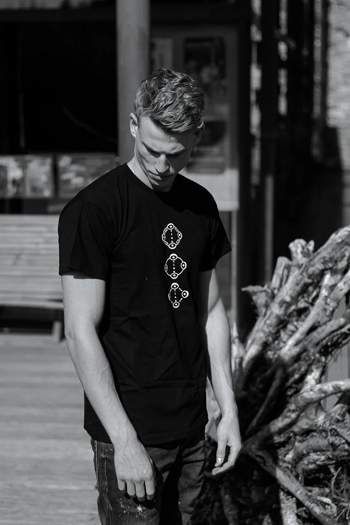 Jeune homme avec des cheveux courts, portant un t-shirt noir avec des dessins de molécules, regardant vers le bas, en extérieur, en noir et blanc.