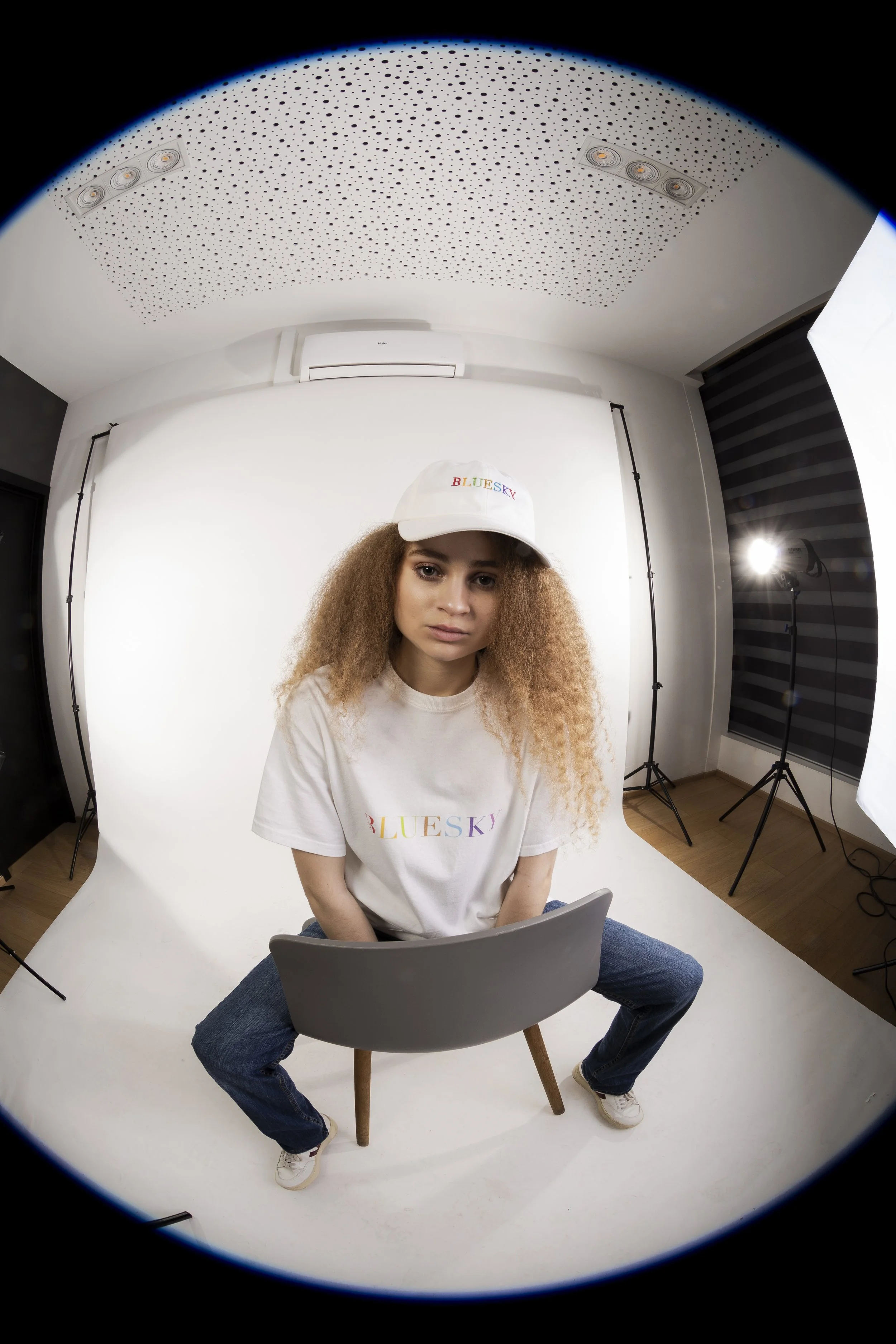 Jeune femme assise devant un fond blanc dans un studio photo, portant une casquette et un t-shirt blanc avec 'BLUESKY' écrit en lettres colorées, et des chaussures blanches.