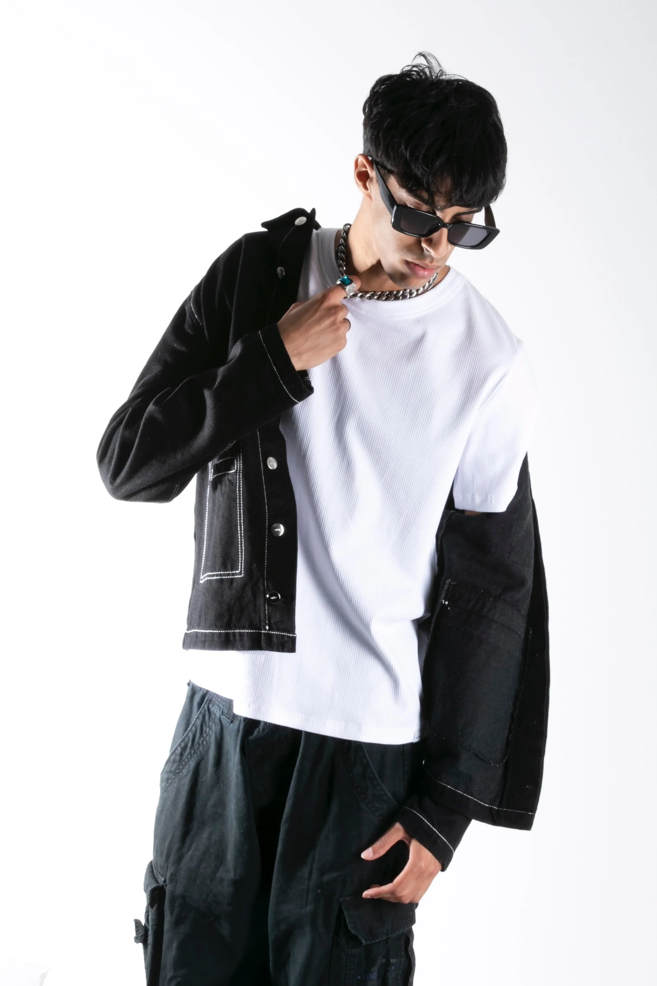 Jeune homme portant des lunettes de soleil, une chaîne en métal, un t-shirt blanc, une veste en jeans noir avec coutures blanches, et un pantalon cargo noir, posant contre un fond blanc.