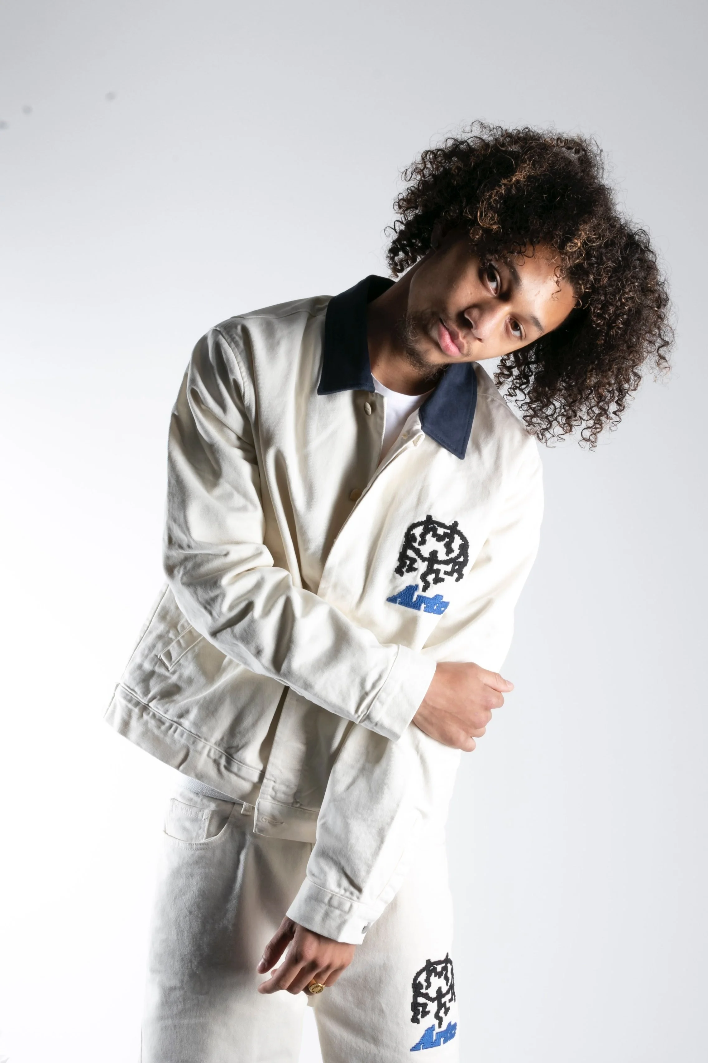 Jeune homme avec des cheveux bouclés portant une veste et un pantalon blancs avec des logos noirs et bleus, posant de profil