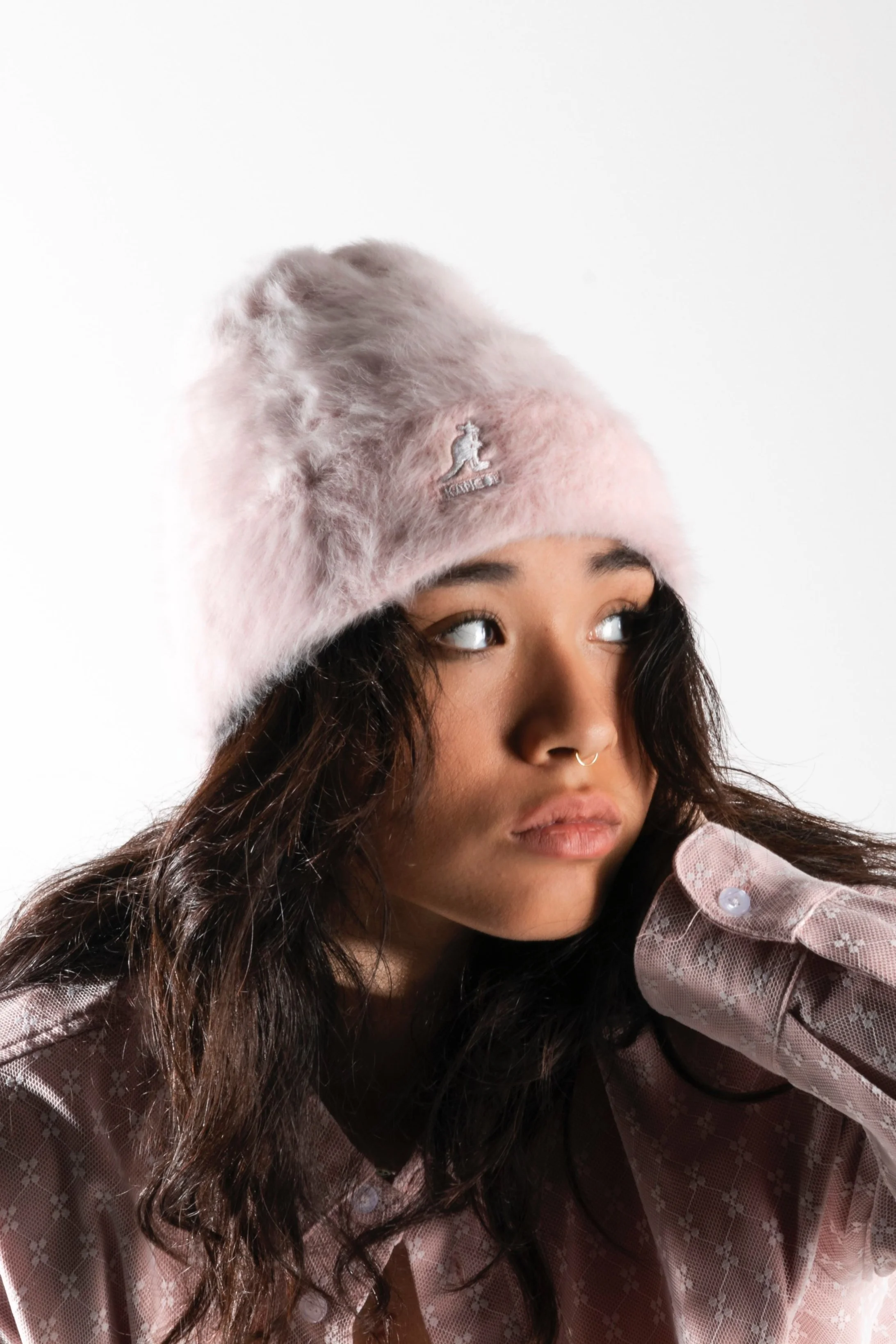 Une jeune femme portant un bonnet en fourrure rose clair et un chemisier rose avec des motifs floraux. Elle a des cheveux bruns ondulés, un nez avec un bijou en septum, et elle regarde vers la droite avec une expression neutre.