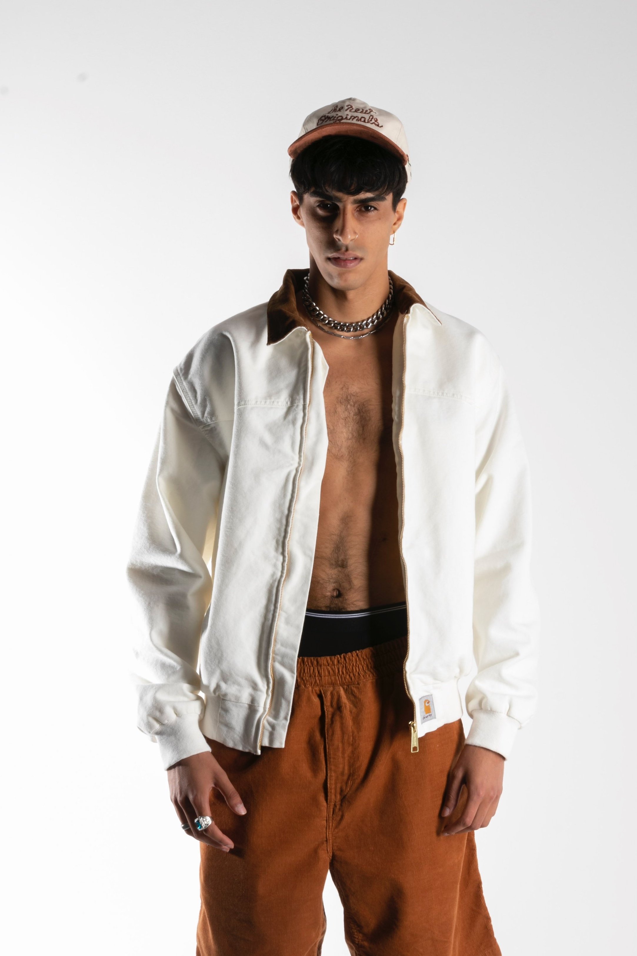 Un homme porte une veste blanche ouverte, un chapeau, des bijoux et un pantalon marron. Il est à la mode et pose devant un fond blanc.