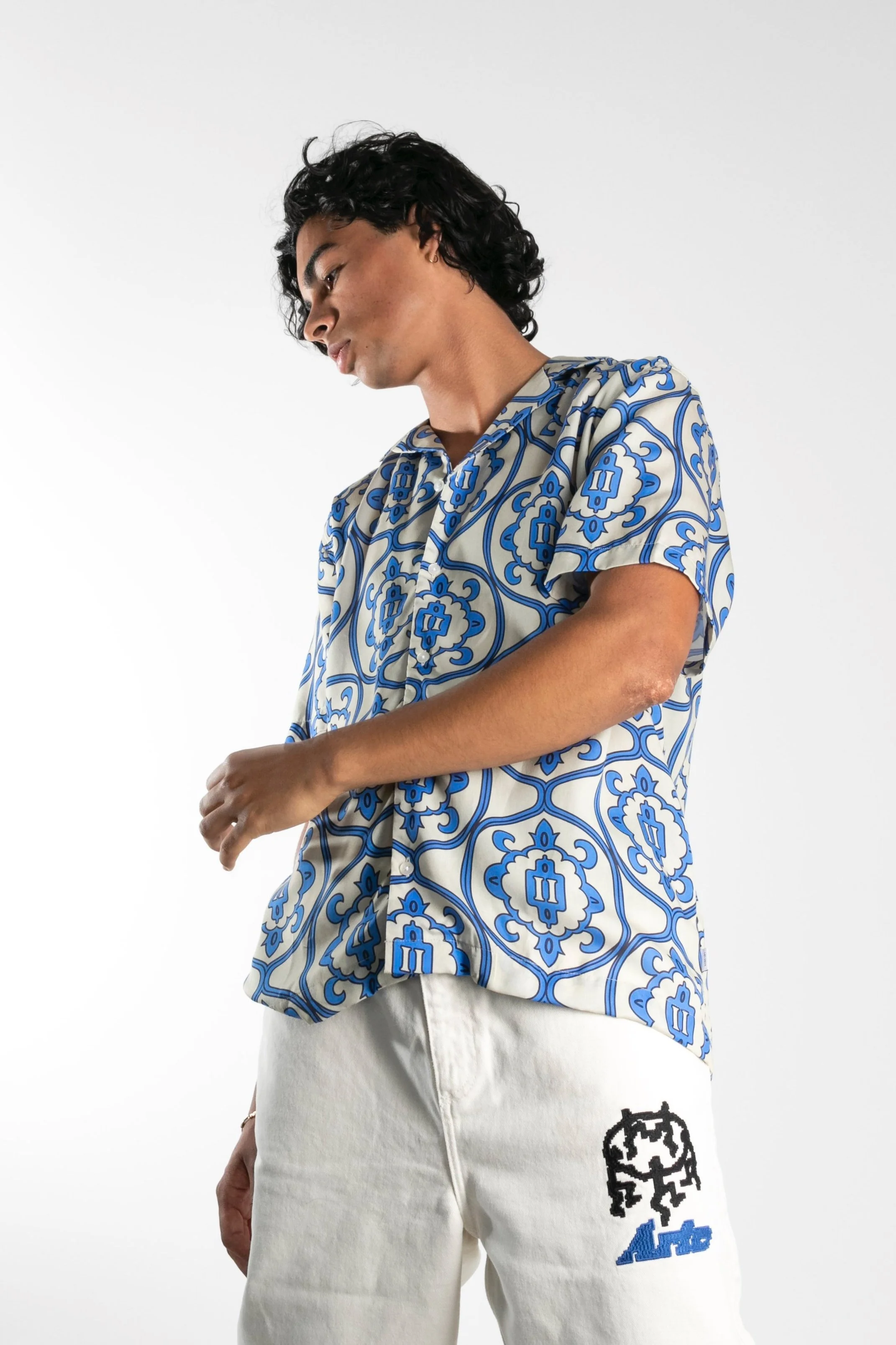 Jeune homme avec des cheveux noirs, portant une chemise courte à motif bleu et blanc, et un pantalon blanc avec un logo noir et bleu.