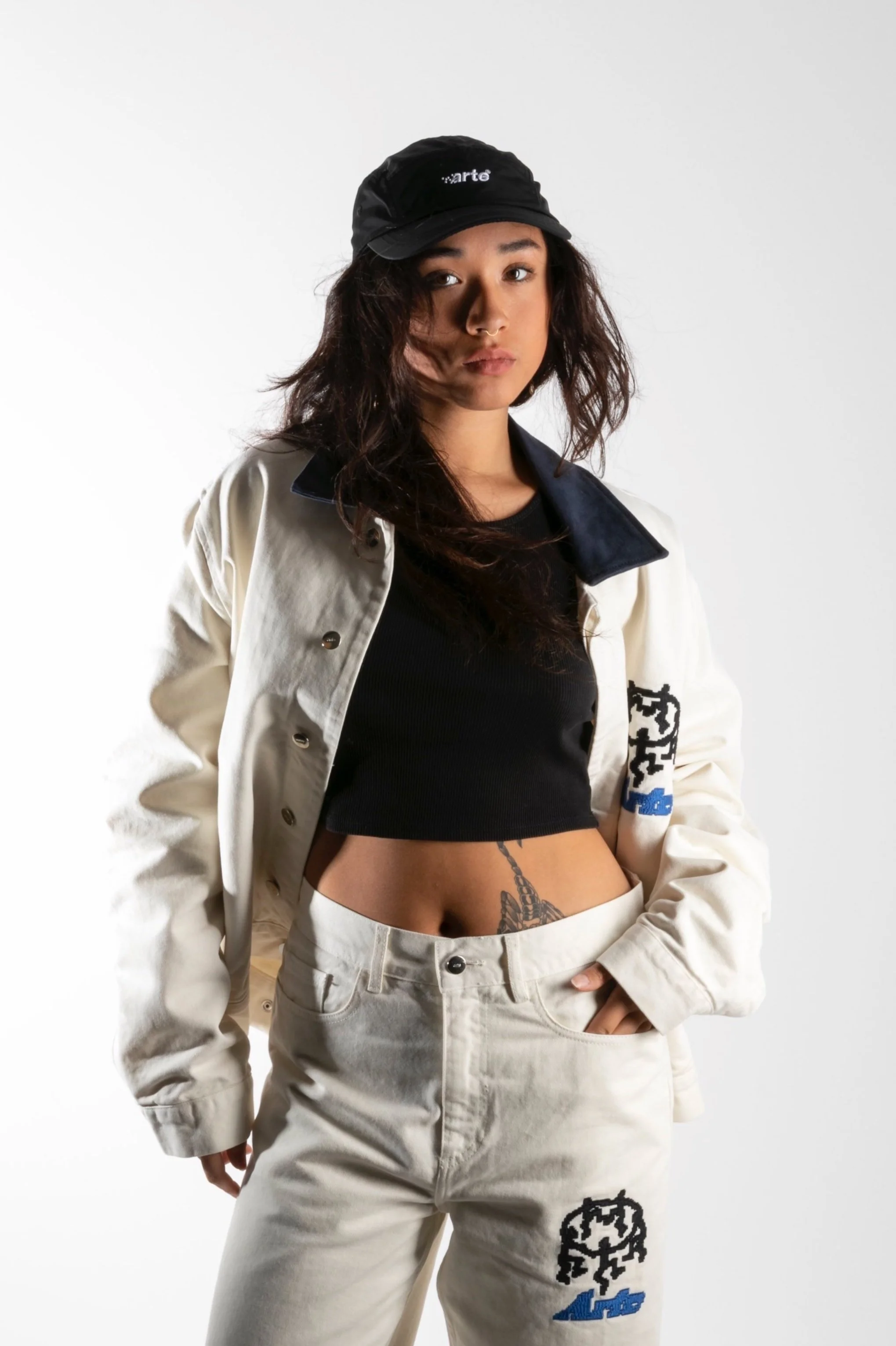 Jeune femme portant une casquette noire, un haut court noir, une veste blanche et un pantalon blanc avec des logos, se tenant contre un fond blanc.