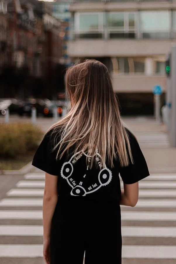 Jeune femme avec des cheveux longs, porte un t-shirt noir avec un design circulaire blanc, se tenant devant un passage pour piétons en ville.