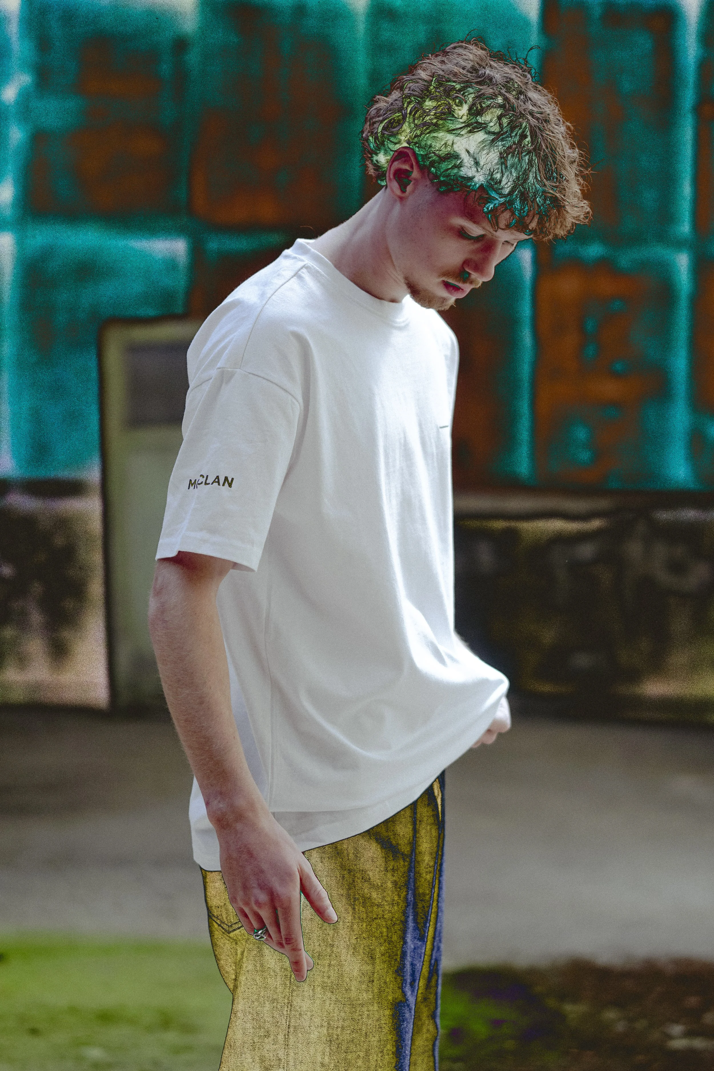Jeune homme aux cheveux bouclés portant un t-shirt blanc et un pantalon jaune avec des motifs bleus, dans un décor coloré et féerique.