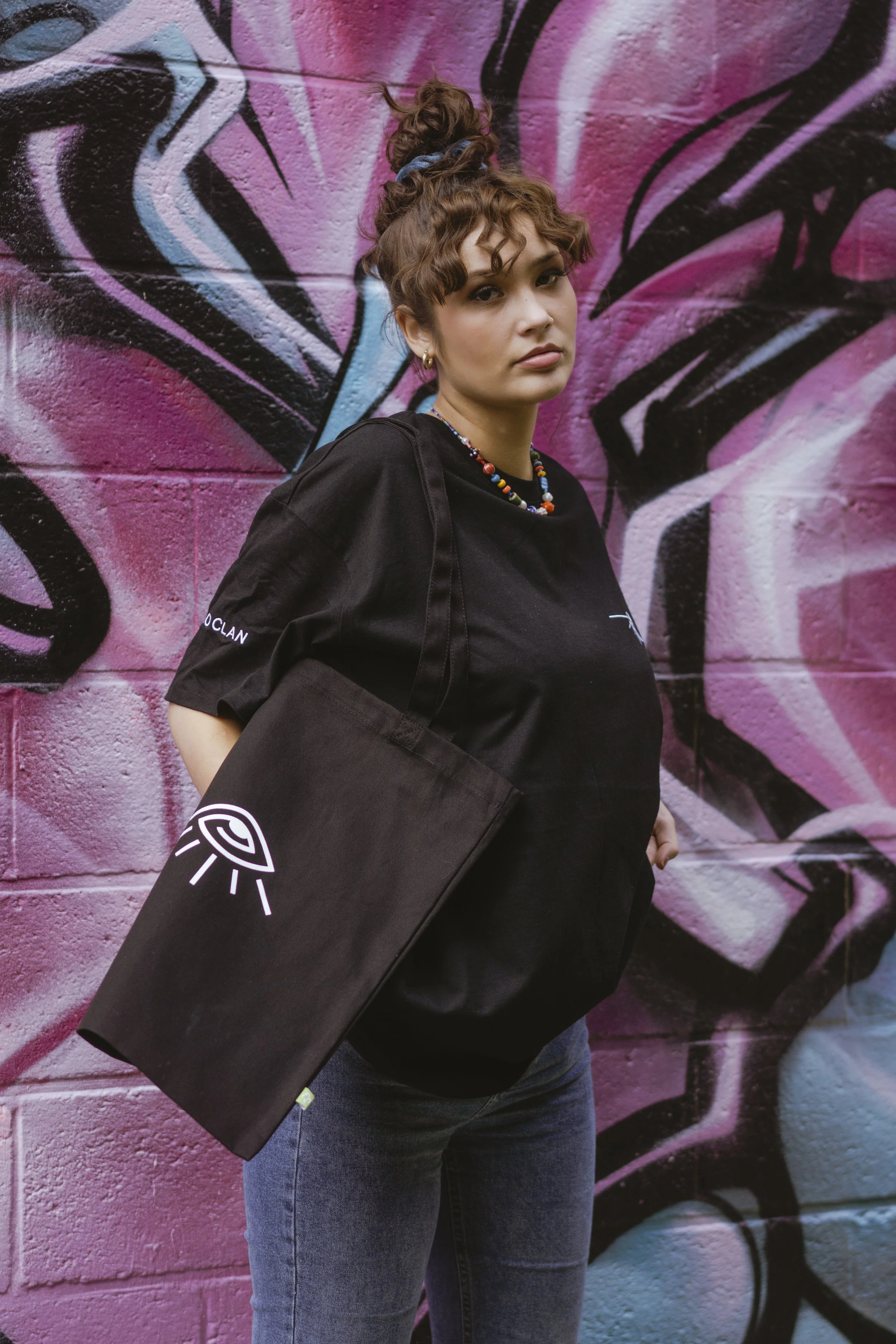 Jeune femme portant un sac noir avec un logo, vêtue d'un t-shirt noir et d'un collier de perles, debout devant un mur en graffiti rose et noir.