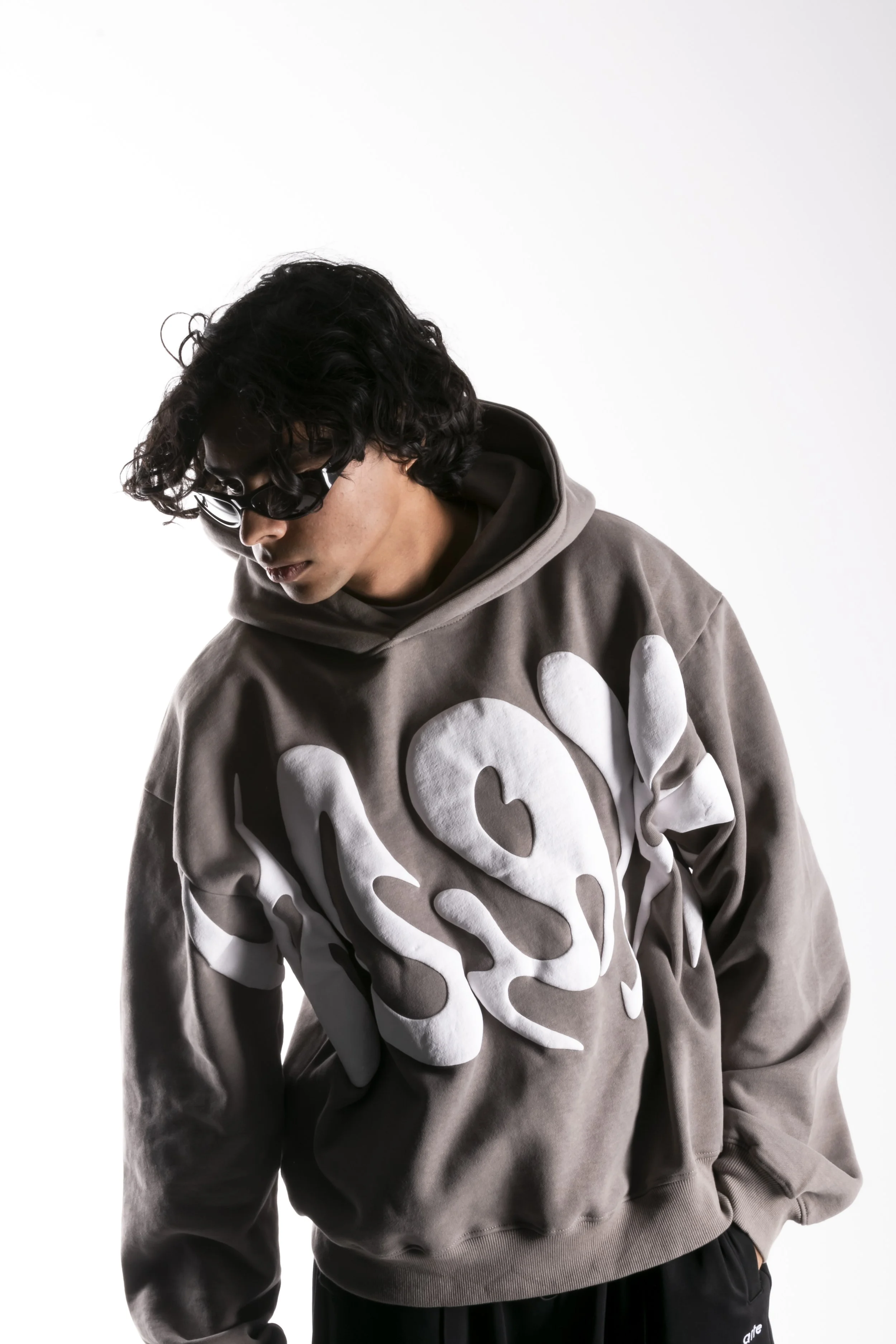 Jeune homme portant un sweat à capuche gris avec un motif blanc en relief, lunettes de soleil noires, regard baissé, fond blanc