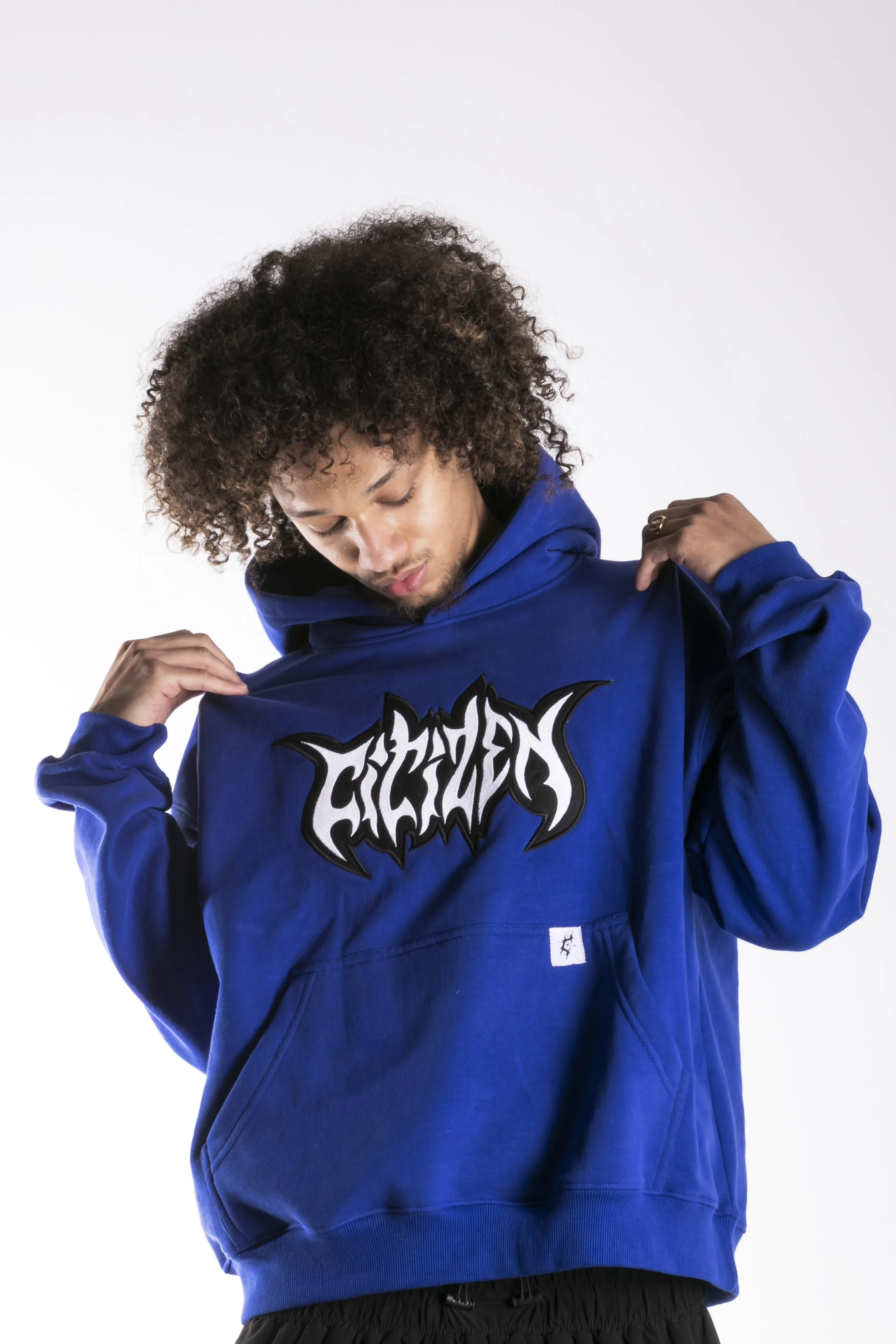Jeune homme portant un sweat à capuche bleu avec un logo blanc et noir, se regardant vers le bas, sur fond blanc.