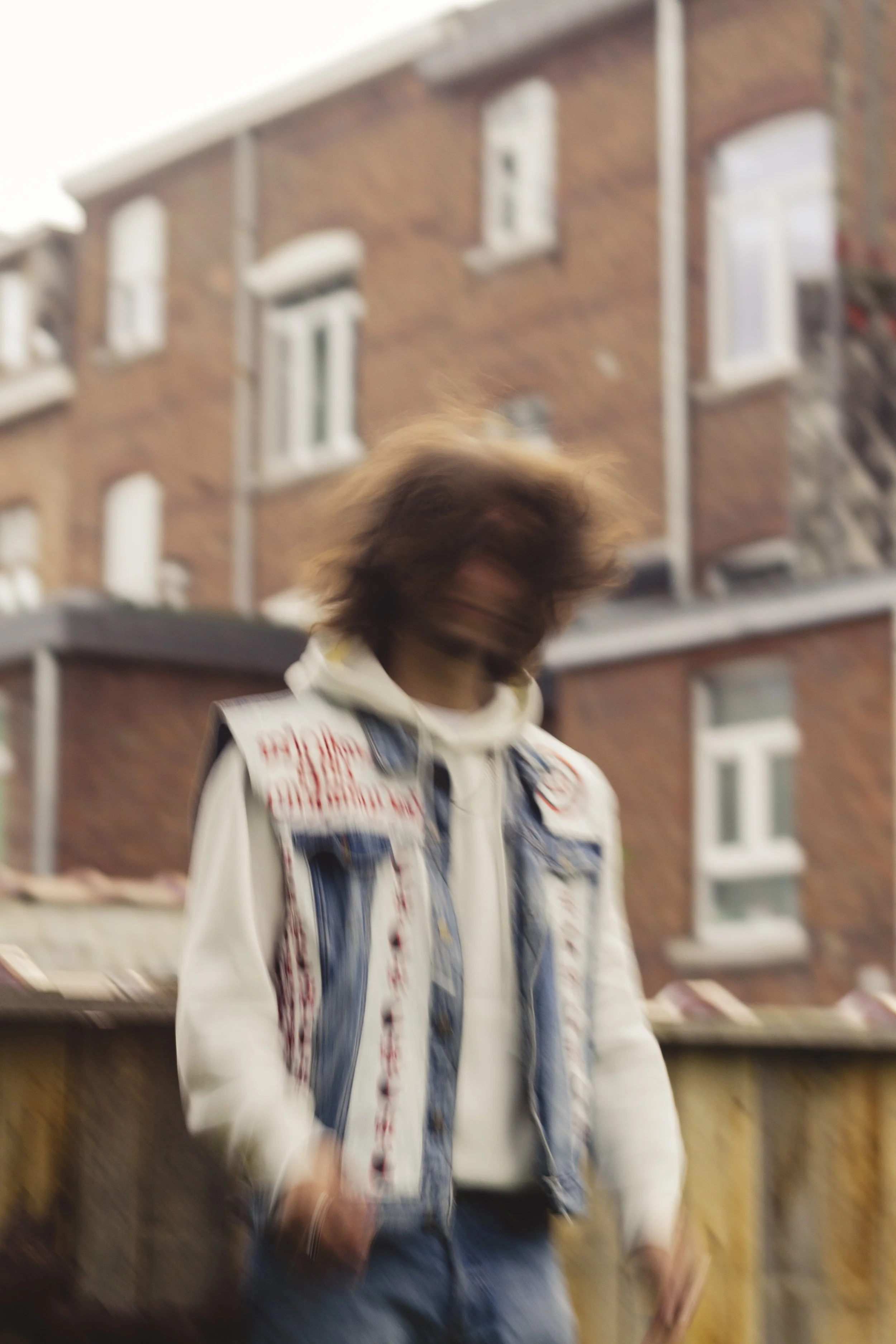 Une personne avec des cheveux longs et bouclés portant une veste en jean et un sweat blanc, debout devant une maison en briques. La photo est floue, ce qui rend difficile d'identifier les détails précis.
