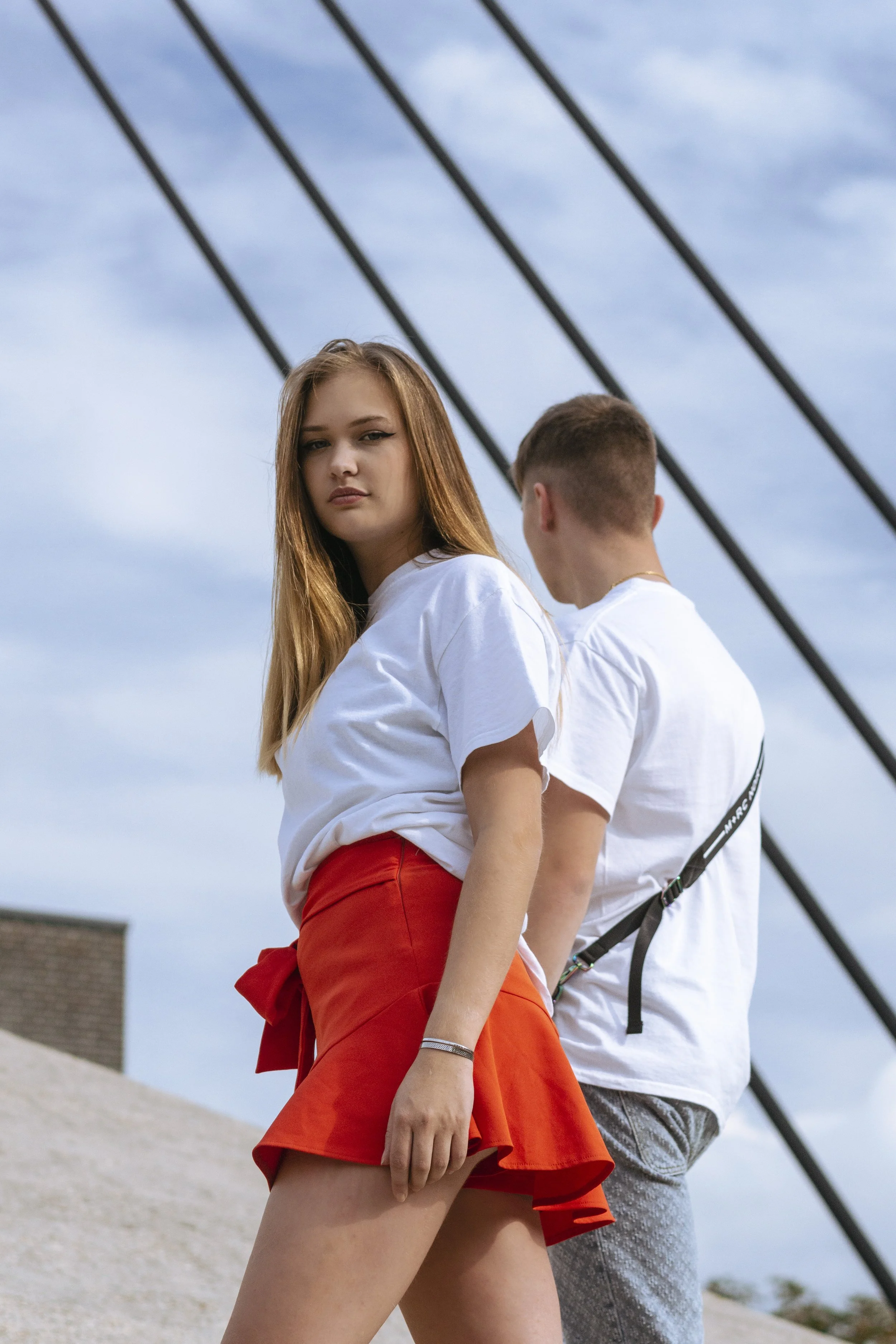 Une jeune femme aux cheveux longs et roux portant une chemise blanche et une jupe courte rouge se tient devant un jeune homme dont le dos est visible, portant un t-shirt blanc et un sac à bandoulière, sous un ciel nuageux.