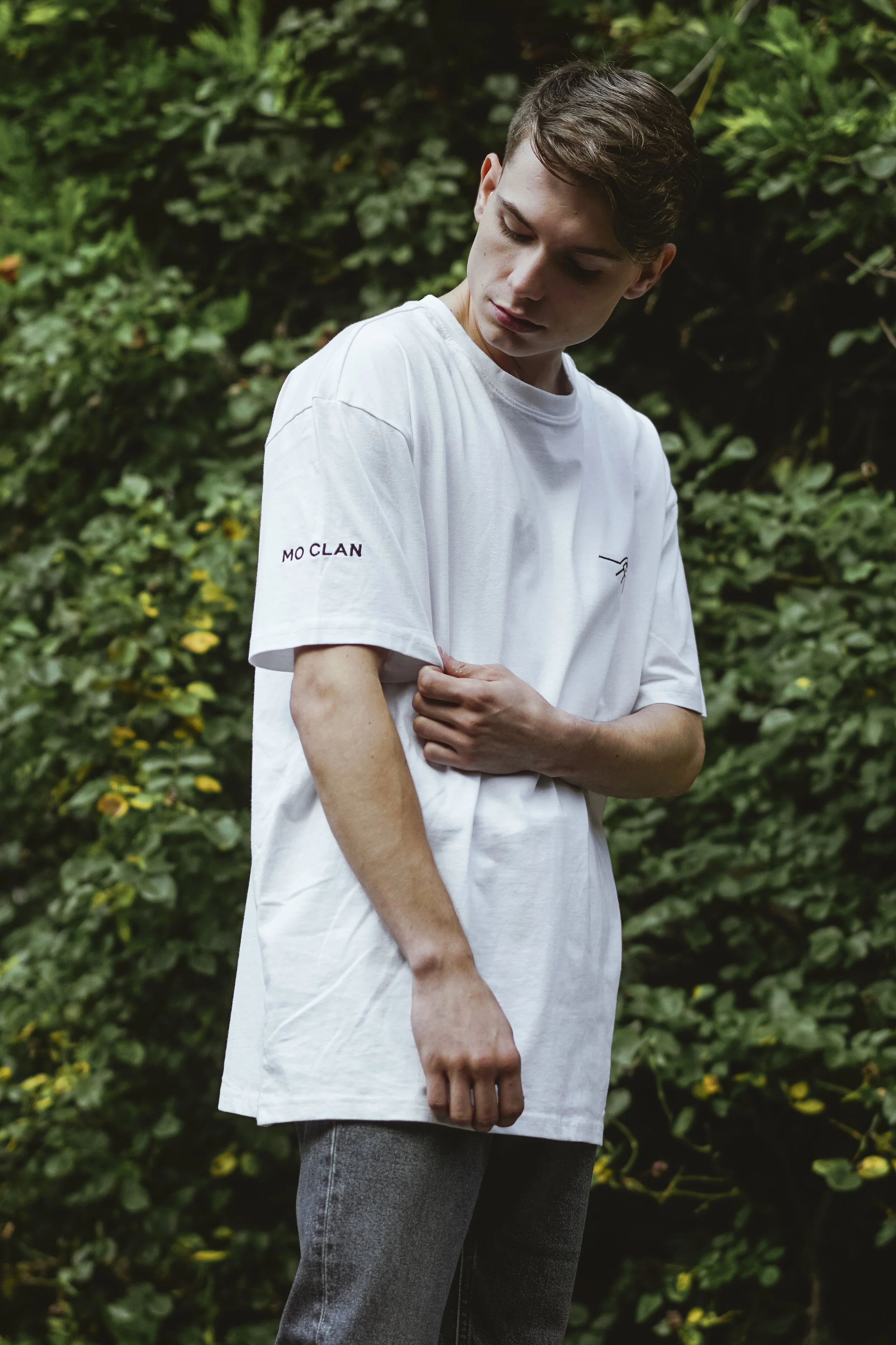 Jeune homme portant un t-shirt blanc et un jean, debout dans la nature, avec des buissons verts en arrière-plan.