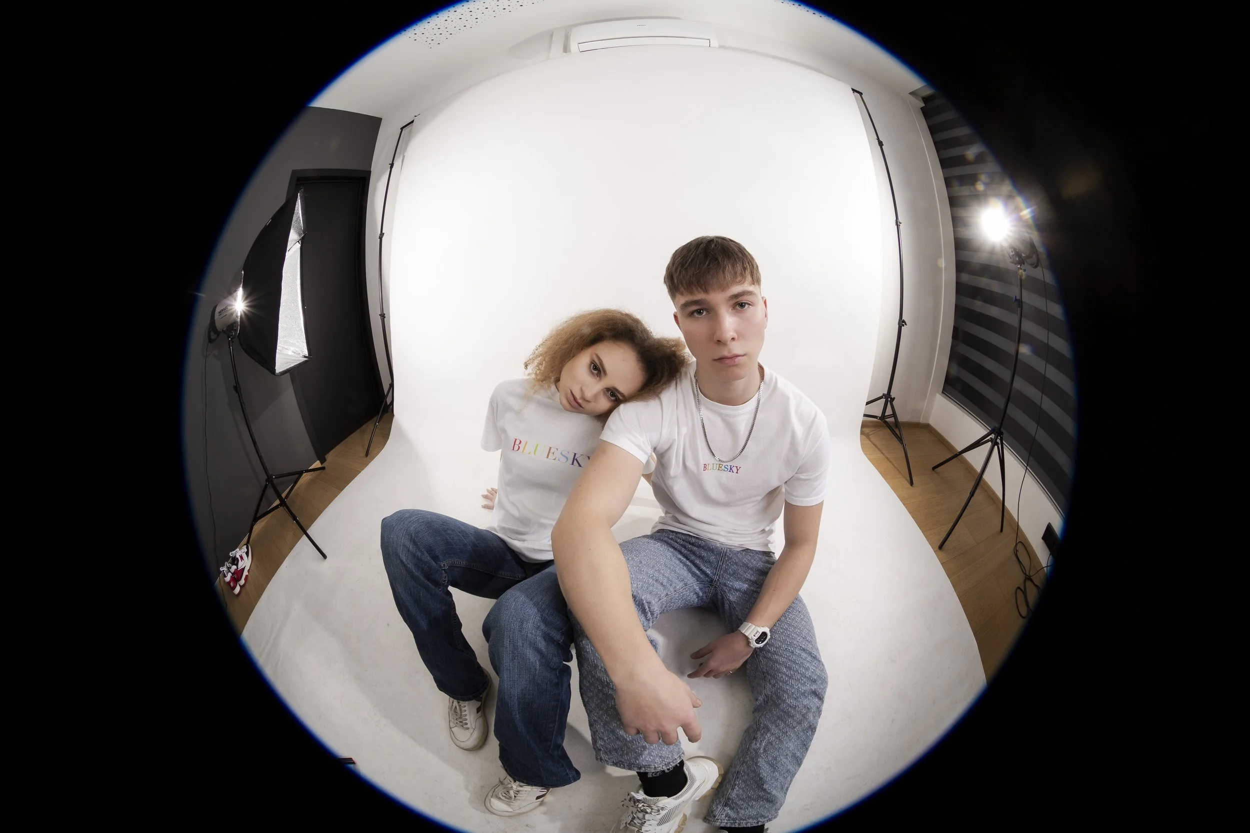 Deux jeunes adultes posent pour une séance photo dans un studio avec un fond blanc, éclairés par deux lampes, portant des t-shirts blancs imprimés 'BLUESKY'