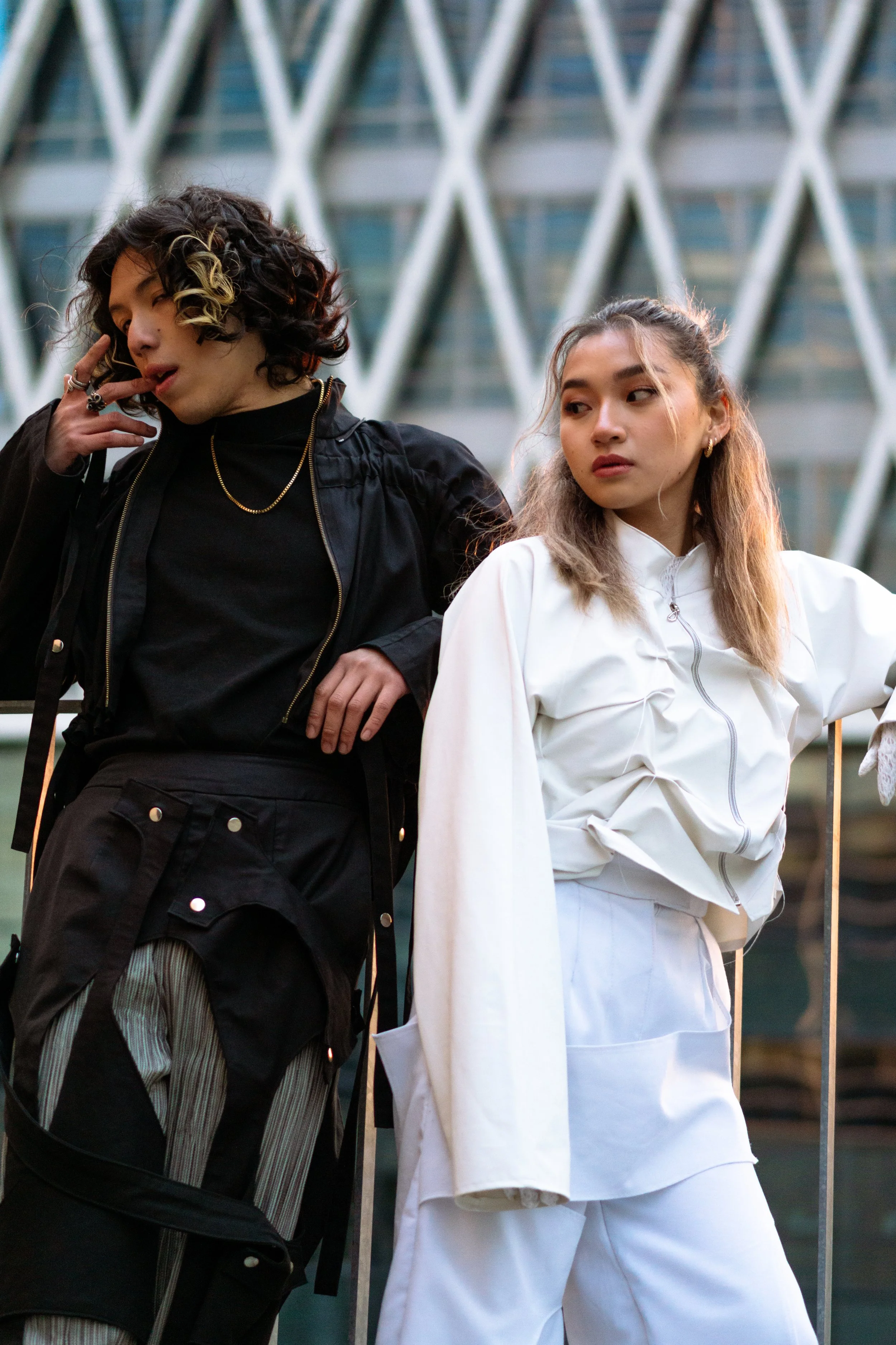 Deux jeunes femmes posant devant un bâtiment moderne, l'une en vêtements noirs et l'autre en blanc, dans une ambiance urbaine.