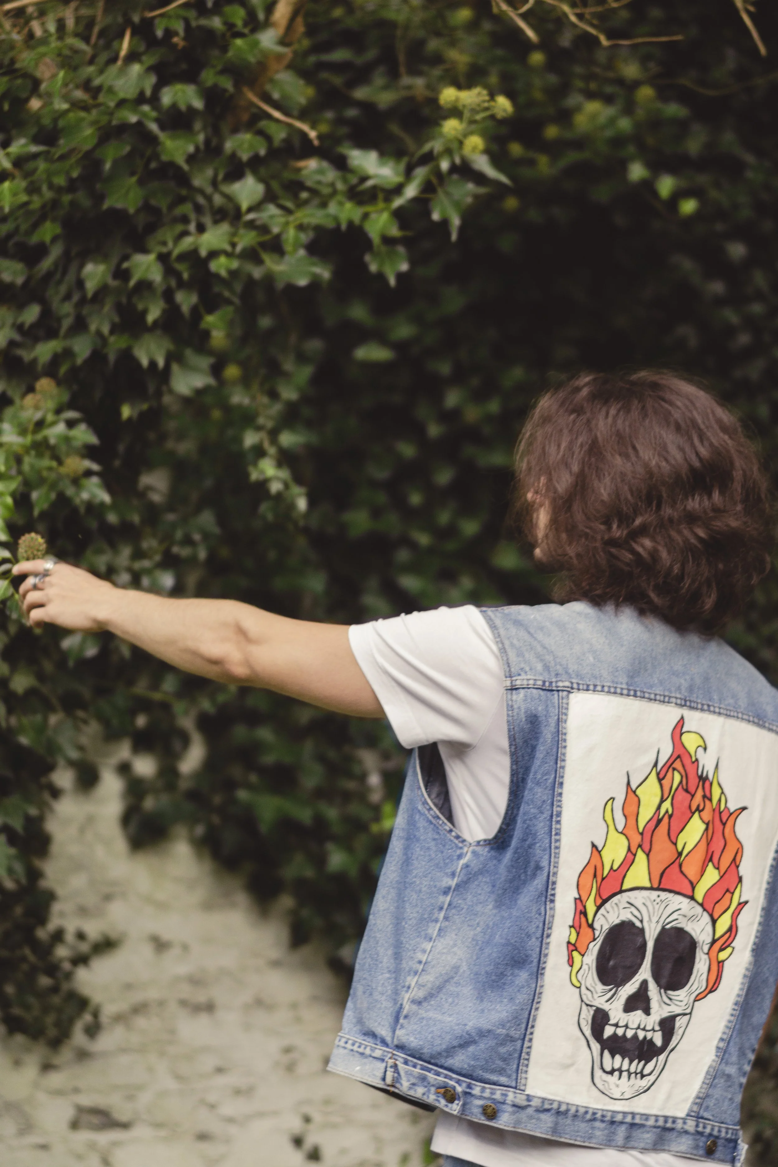 Une personne vue de dos portant un jean sans manches avec une patch d'un crâne en feu, levant la main vers un buisson vert dense.