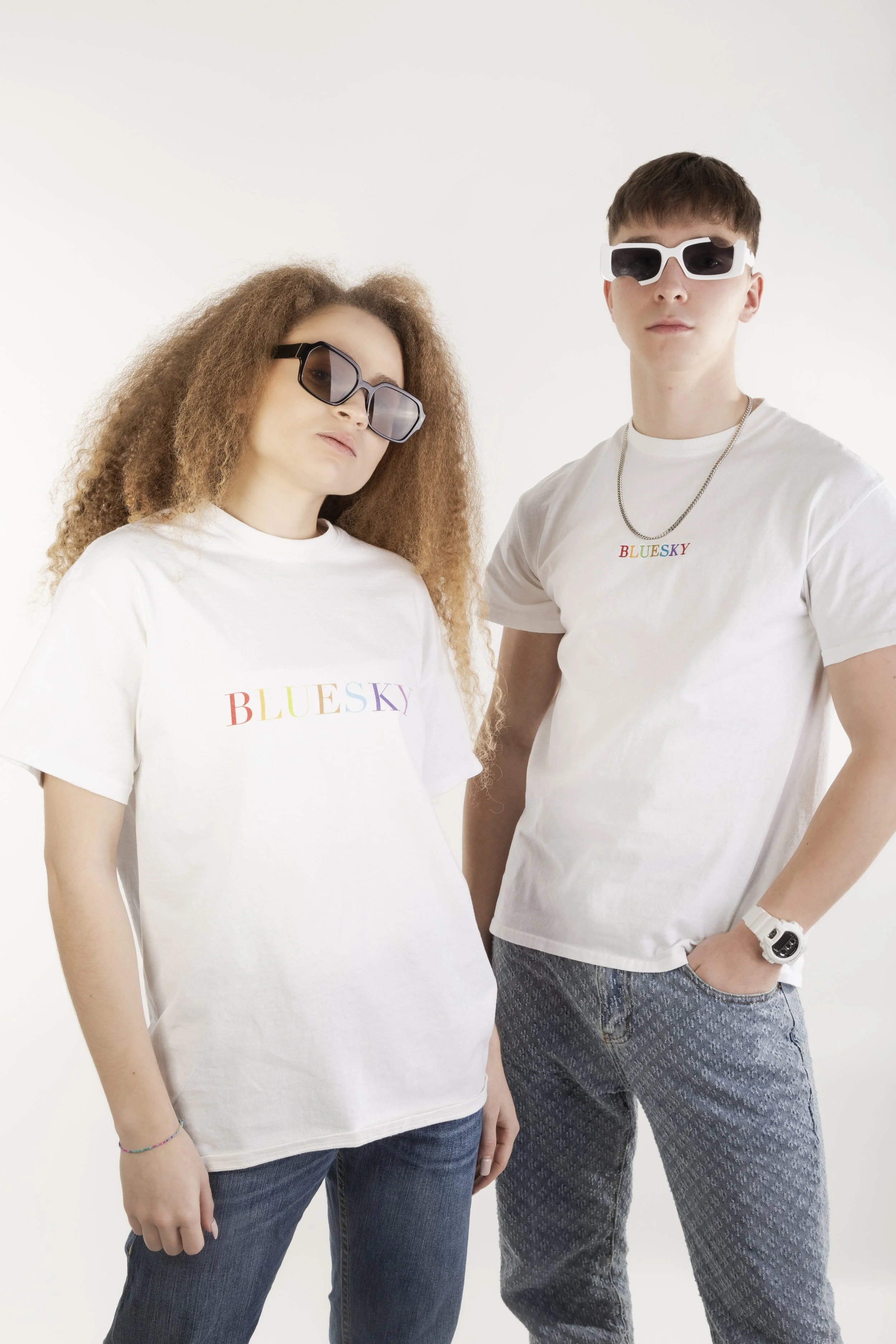 Deux jeunes portant des T-shirts blancs avec le texte 'BLUESKY' arc-en-ciel, portant des lunettes de soleil modernes, posant contre un fond blanc.