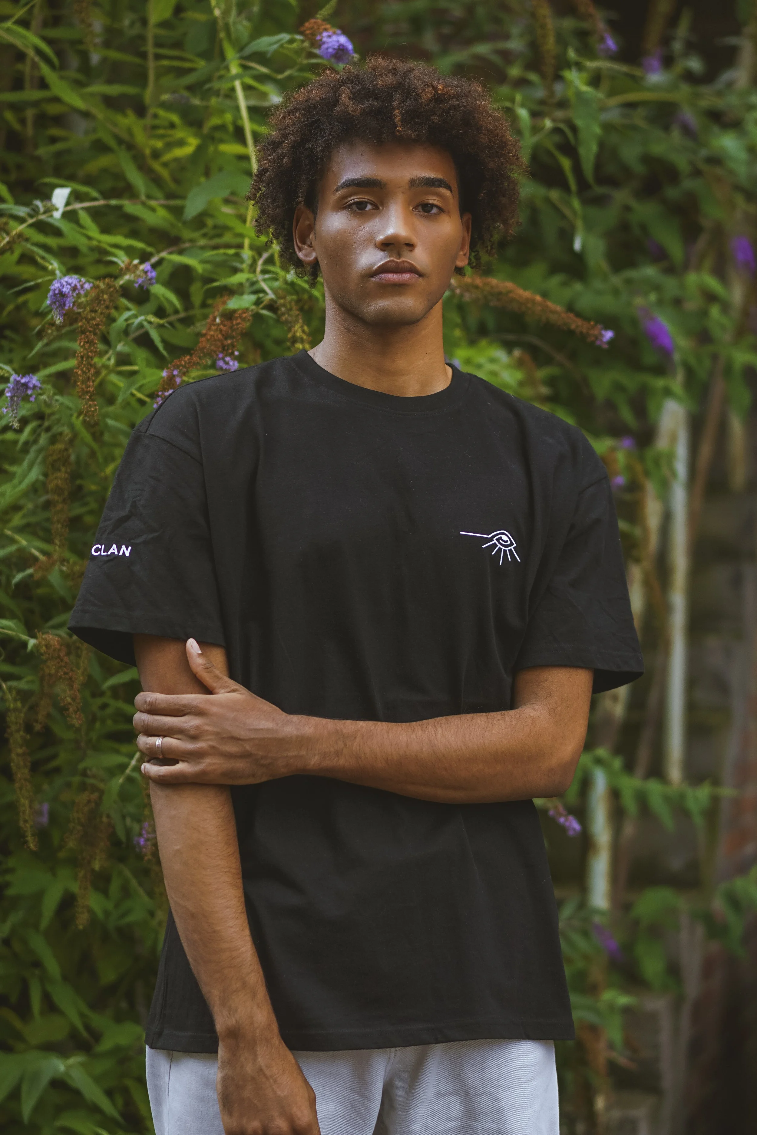 Un jeune homme avec des cheveux bouclés, portant un t-shirt noir avec un dessin minimaliste d'un œil et une inscription "CLAN" sur la manche, se tenant devant une végétation luxuriante avec des fleurs violettes.