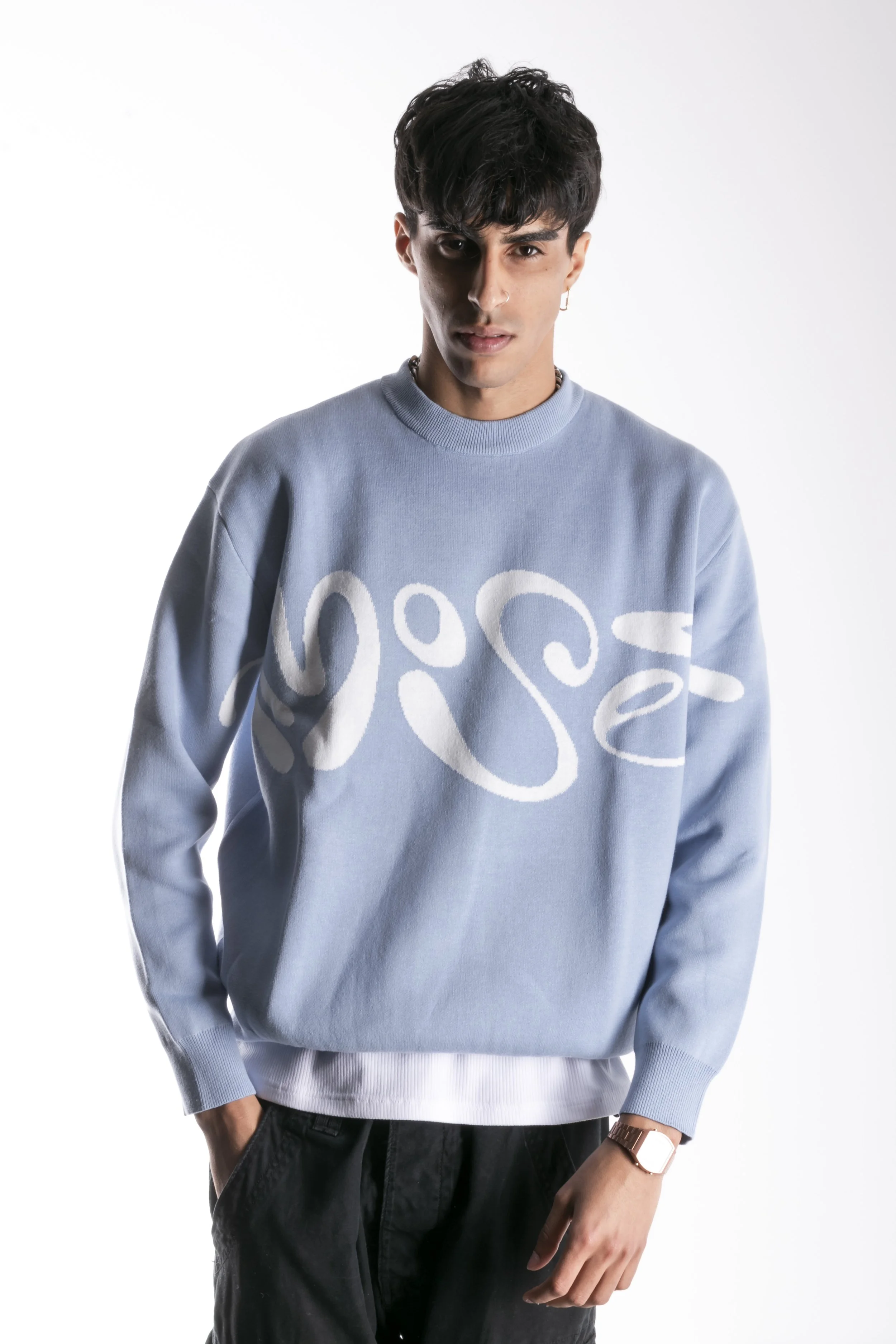 Jeune homme portant un sweat bleu clair avec un motif blanc abstrait, tenant une main dans sa poche, fond blanc.