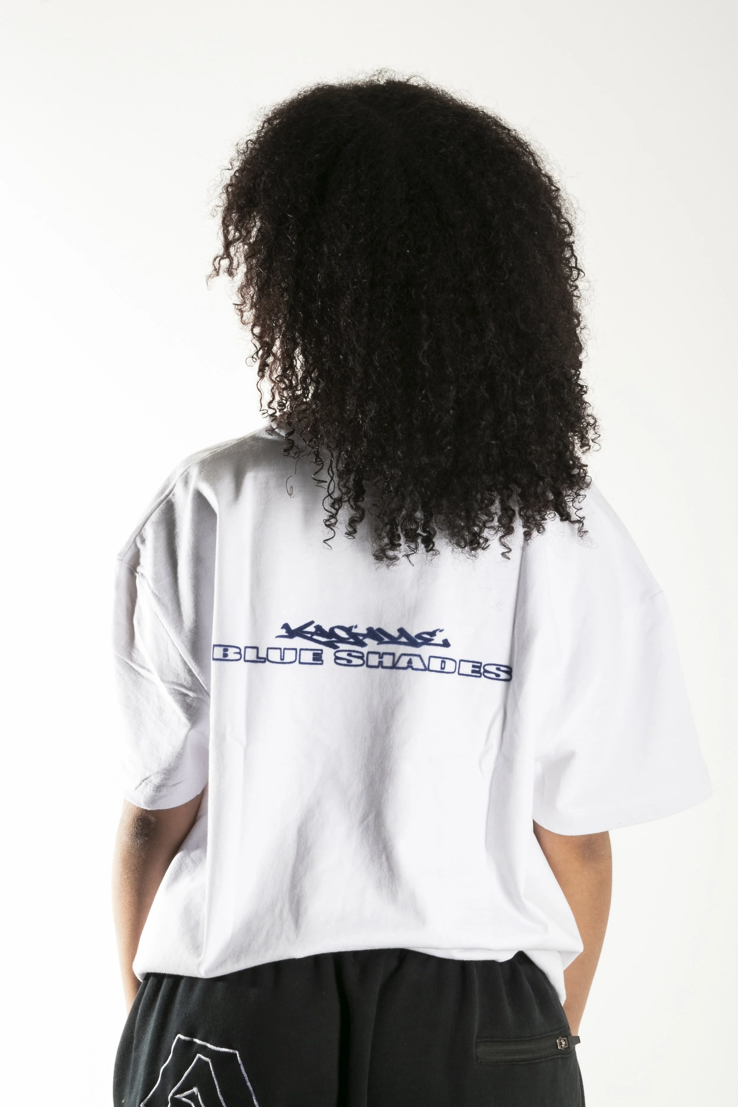 Une personne avec des cheveux bouclés noirs portant un T-shirt blanc avec écrit 'KICKS' et 'BLUE SQUAD' en bleu, et un pantalon noir.