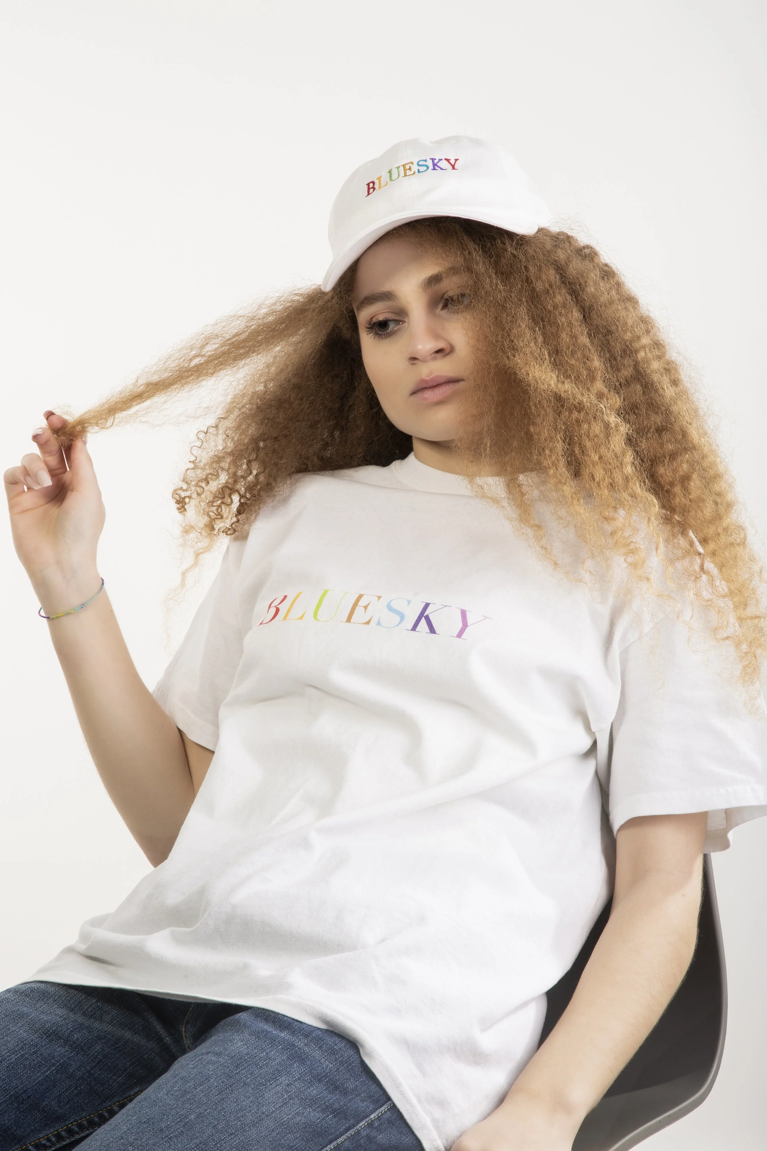 Jeune femme aux cheveux bouclés, portant un t-shirt blanc et une casquette blanche, tous deux avec le mot 'BLUESKY' écrit en lettres colorées, assise sur une chaise, regardant pensivement.