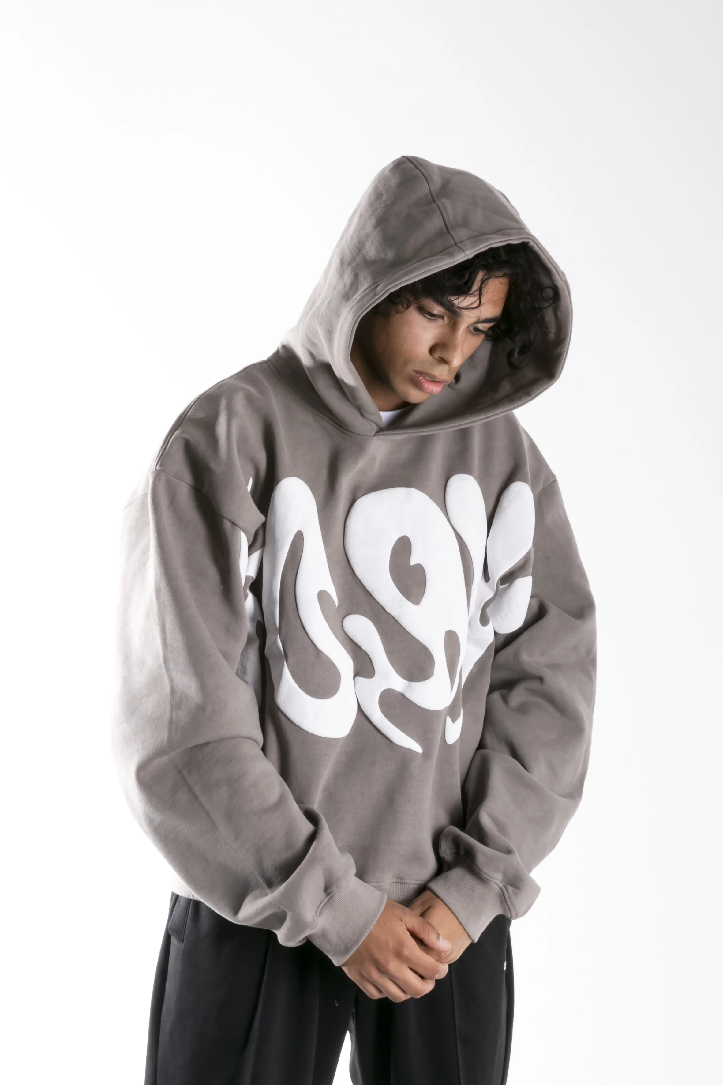 Jeune homme portant un sweat à capuche gris clair avec un motif blanc abstrait et un capuchon, regardant vers le bas, avec les mains jointes devant lui, fond blanc.