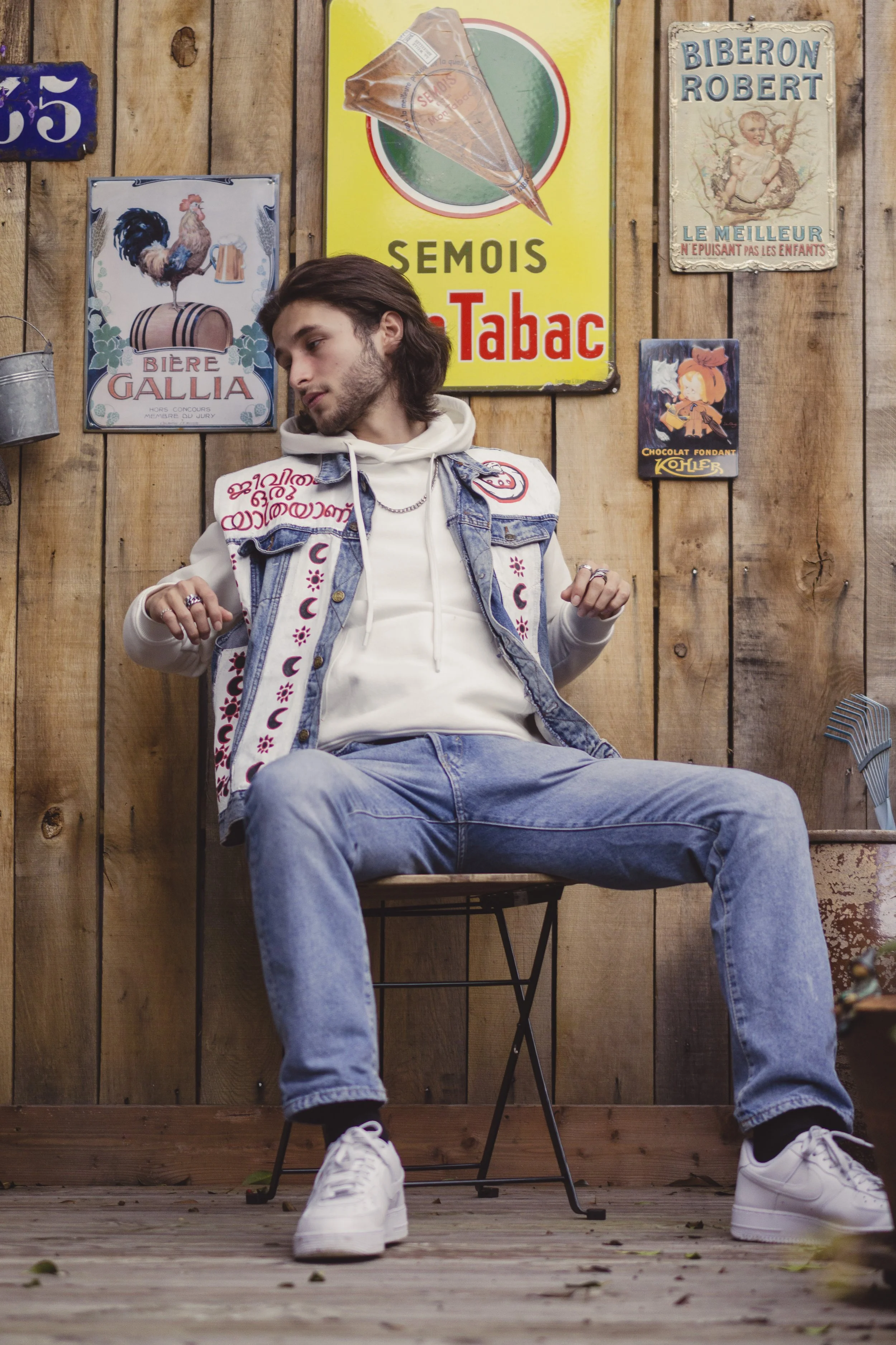 Un jeune homme assis sur une chaise en bois, portant un sweat à capuche blanc, un jean et une veste en jean décorée de motifs de lunes et de fleurs, dans un décor rustique avec des affiches vintage en arrière-plan.